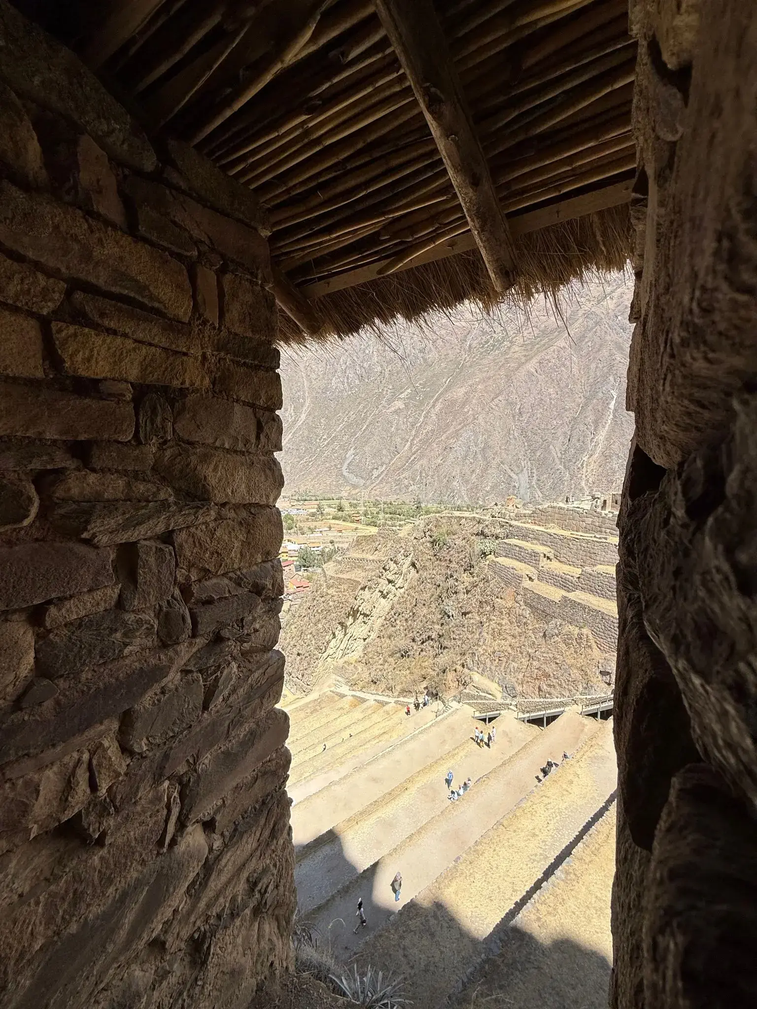 Stone-framed viewpoint looking over terraced slopes of Ollantaytambo in the Sacred Valley, Peru, with small groups of visitors on the paths.