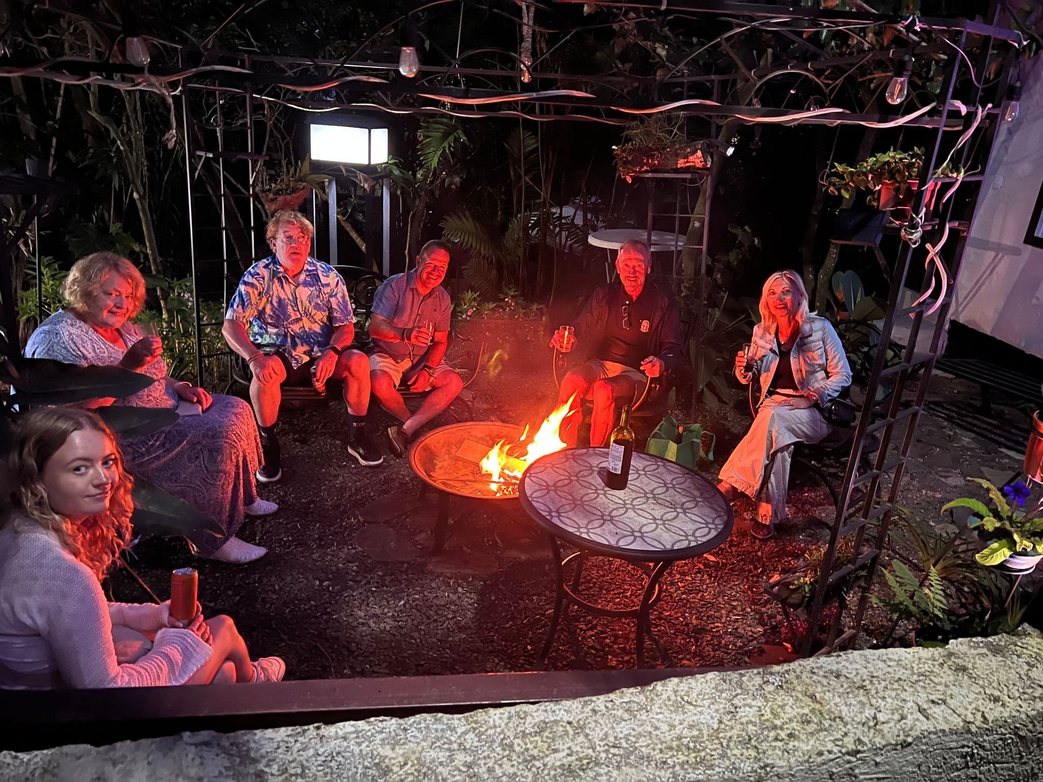 Group of six people sitting around a fire pit at night holding drinks in a tropical backyard, Costa Rica.