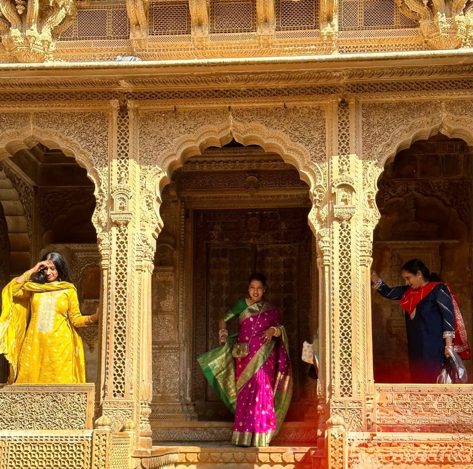 Carved sandstone arches of Patwon Ki Haveli in Jaisalmer framing three women in colorful saris on the steps, India
