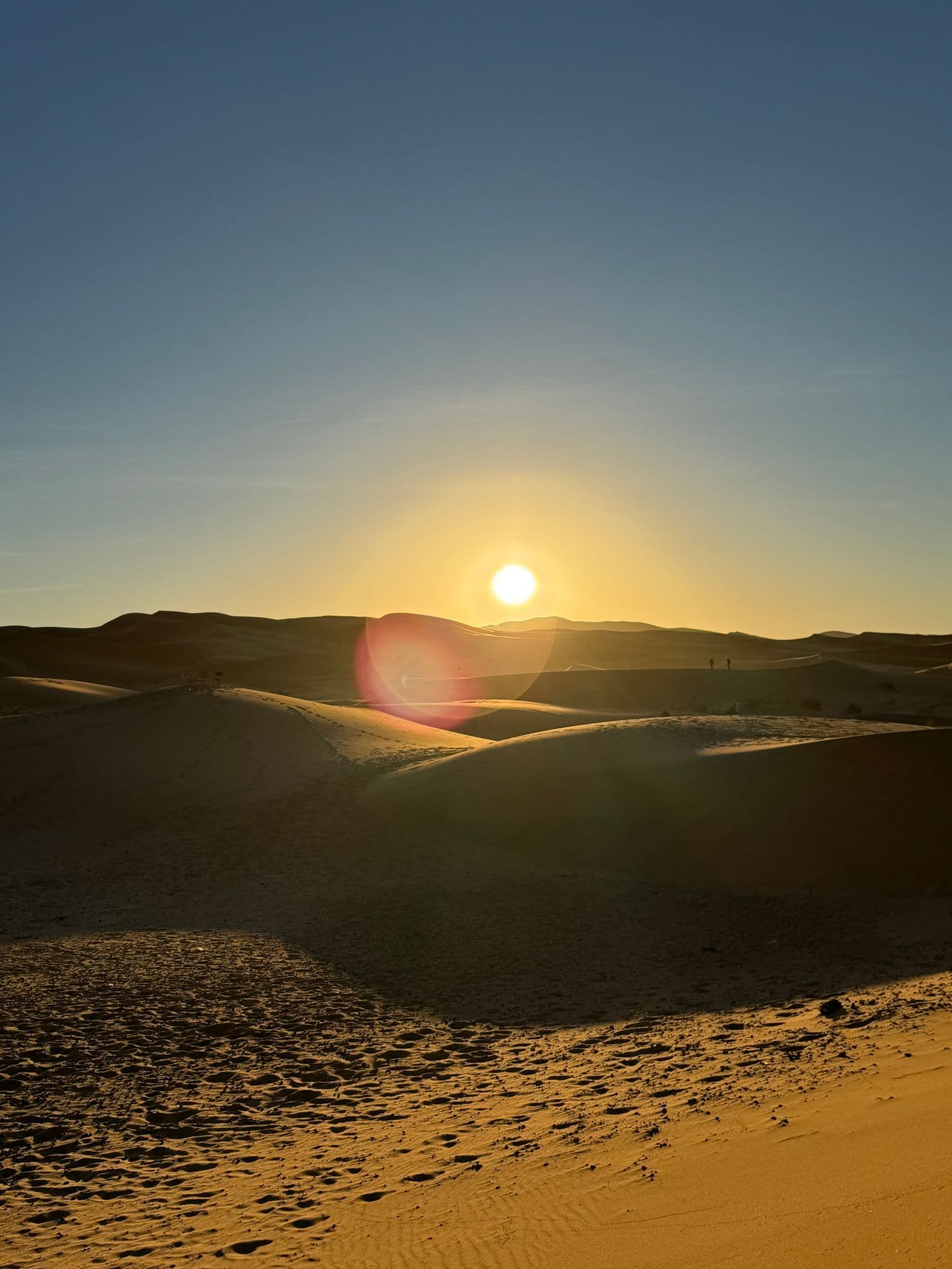 Sunrise over sand dunes in the Sahara Desert with two small people on a distant ridge and footprints in the sand.