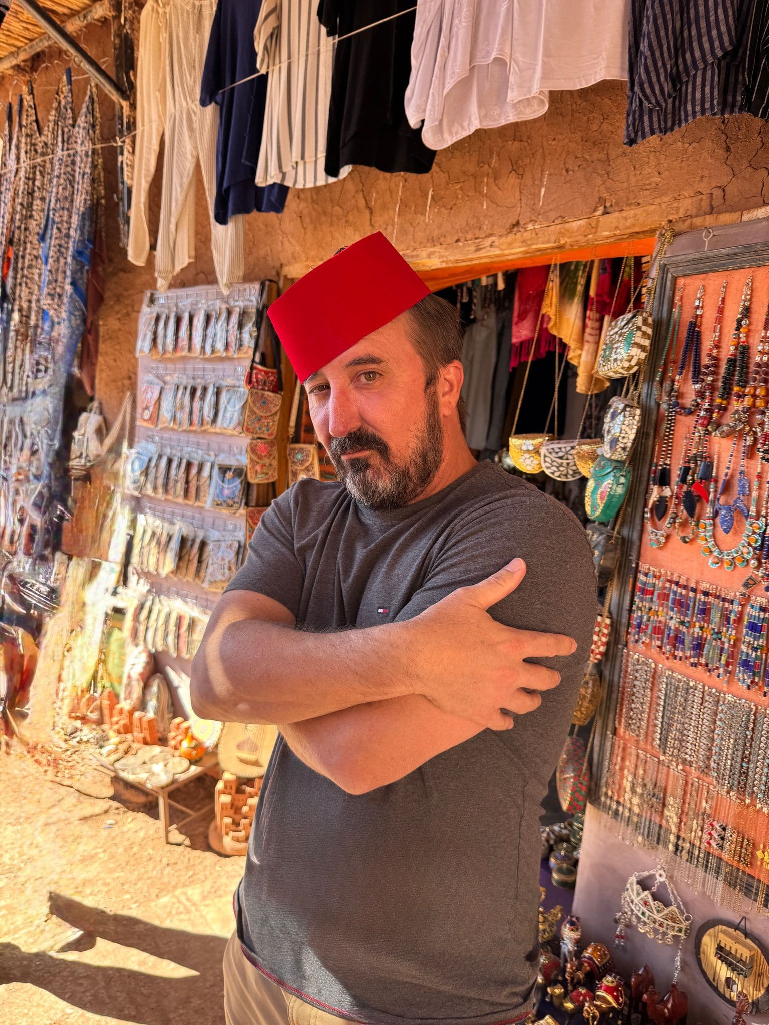 Man wearing a red fez standing with crossed arms in a busy souk stall displaying jewelry and textiles, Marrakech, Morocco.