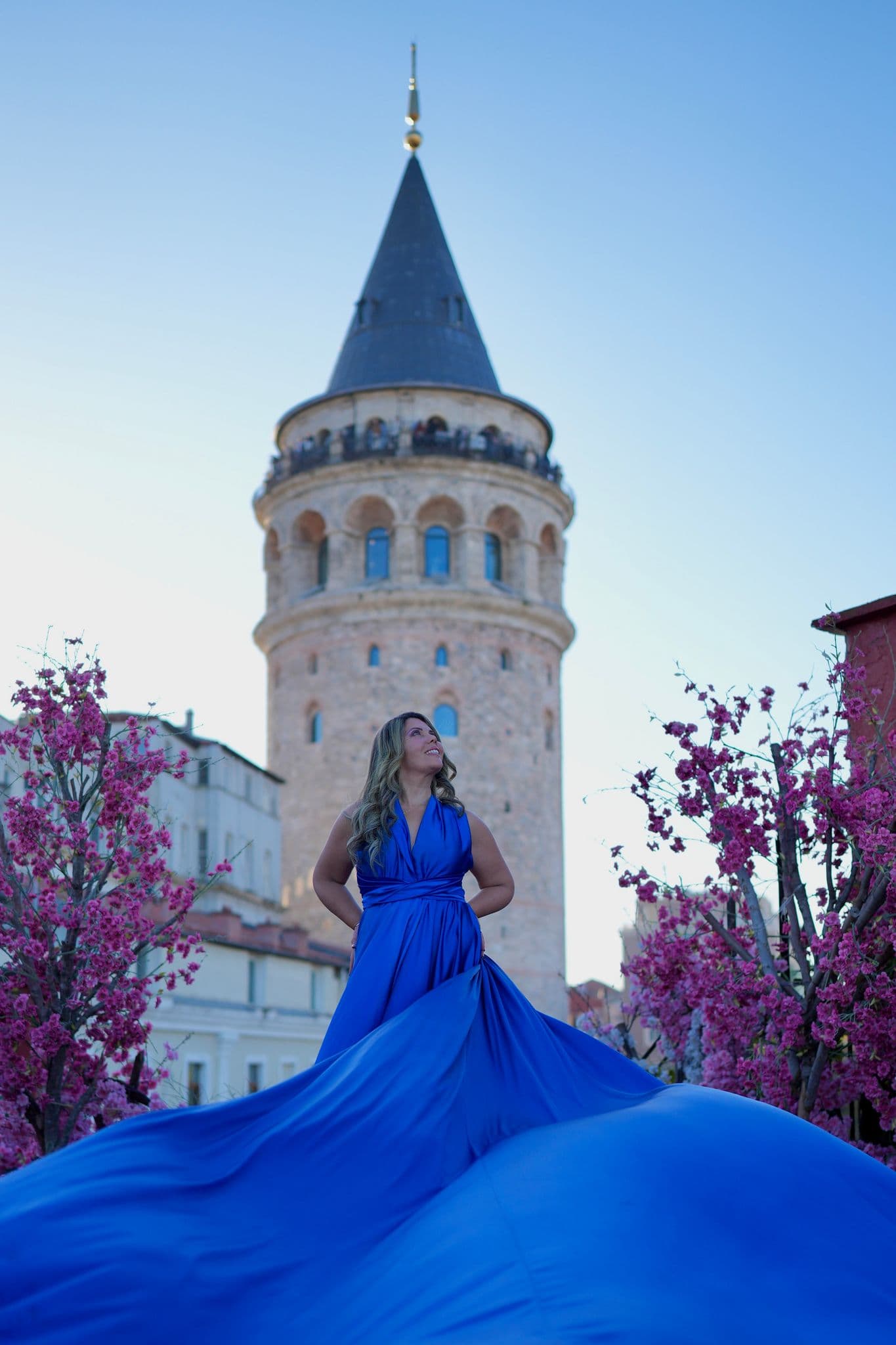 Galata Tower rising behind a woman in a flowing blue dress standing on a rooftop framed by pink blossom trees, Istanbul, Turkey.