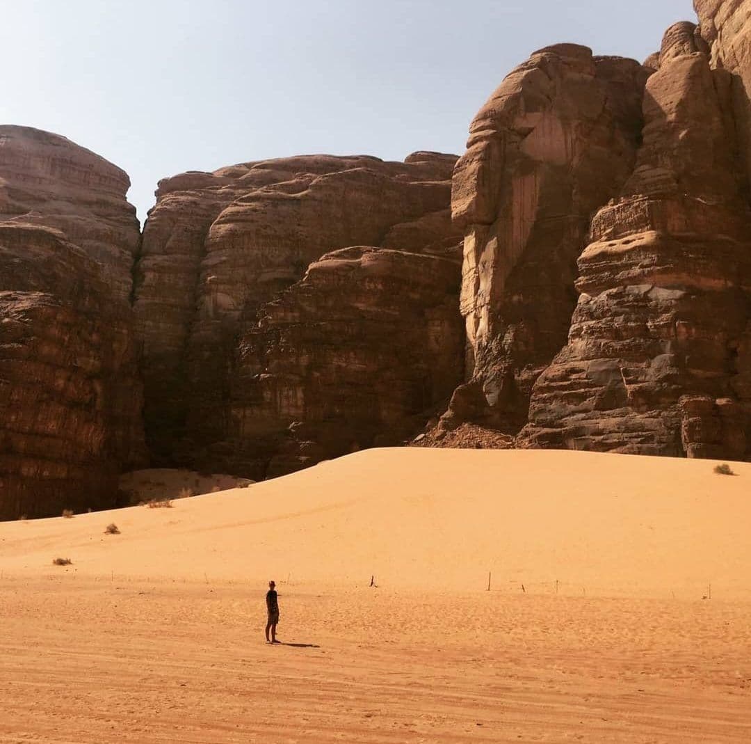 Sandstone cliffs at Wadi Rum, Jordan with a lone traveler standing on a wide sand dune in the desert.