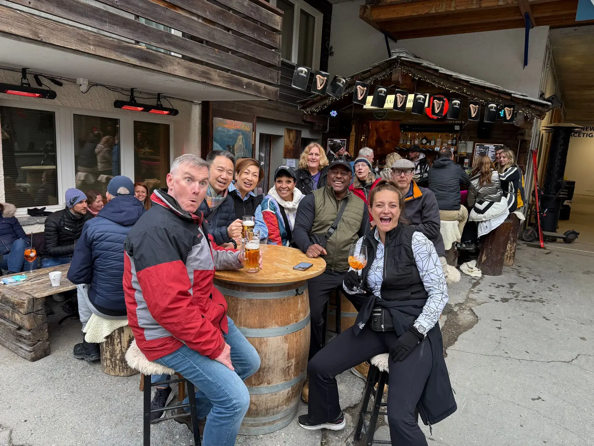 Group of travelers holding drinks around a wooden barrel table outside an alpine bar in Zermatt, Switzerland.