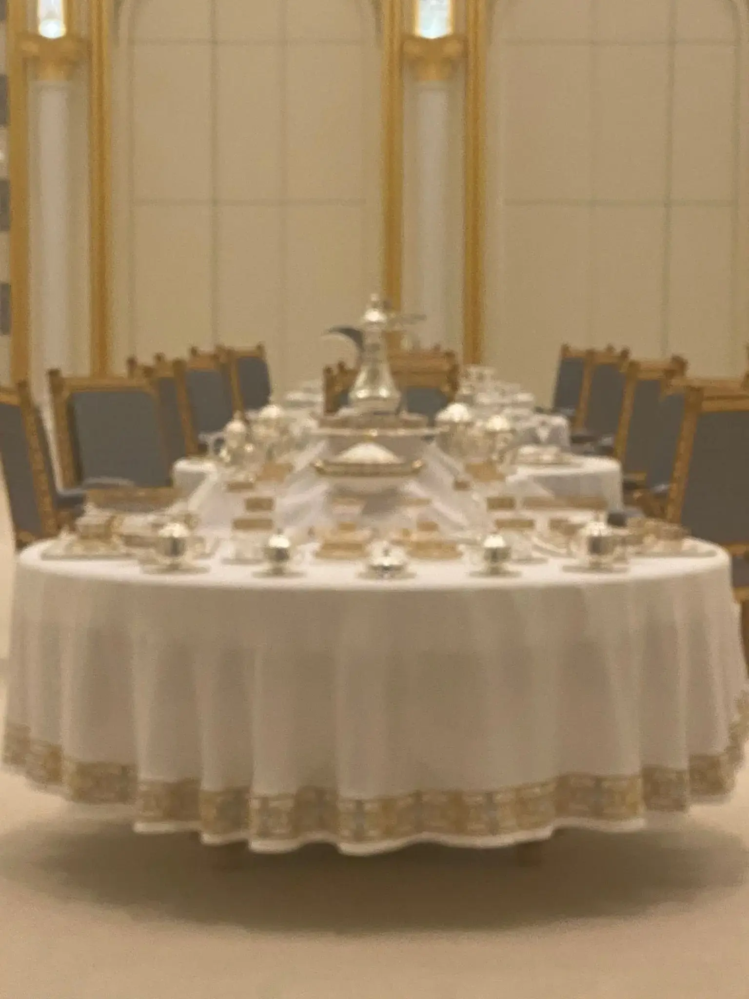Round formal dining table set with silver service in an ornate palace dining room, gilded chairs and paneling visible.