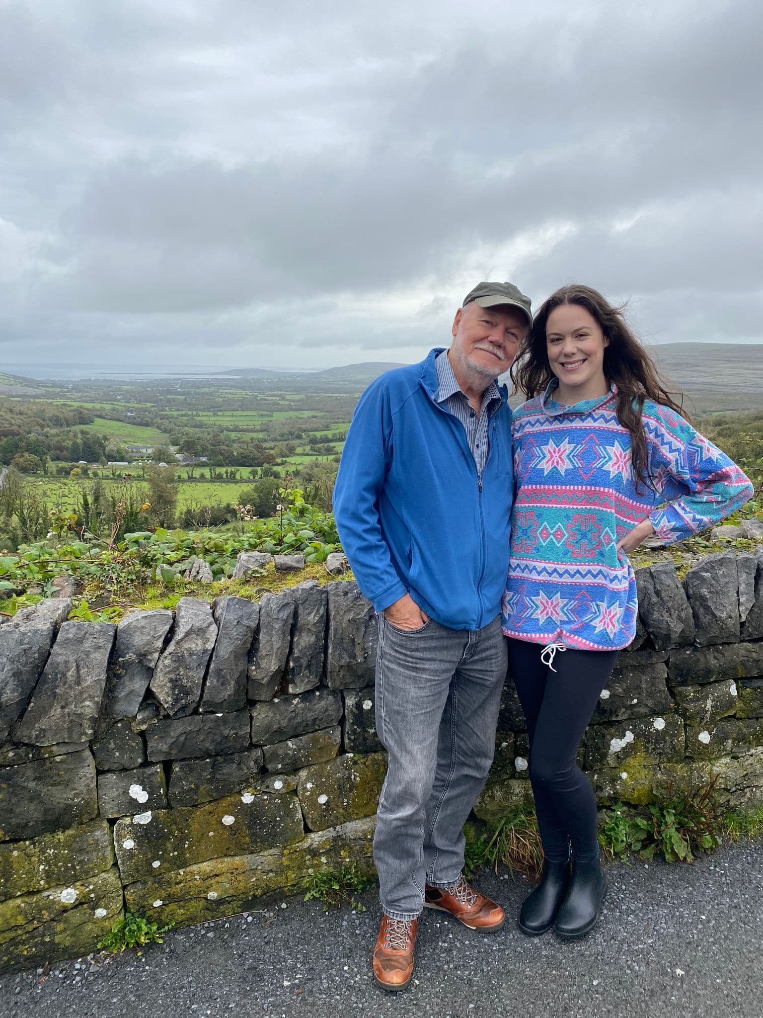 Two travelers standing by a stone wall overlooking green fields and distant coastline in rural Ireland.