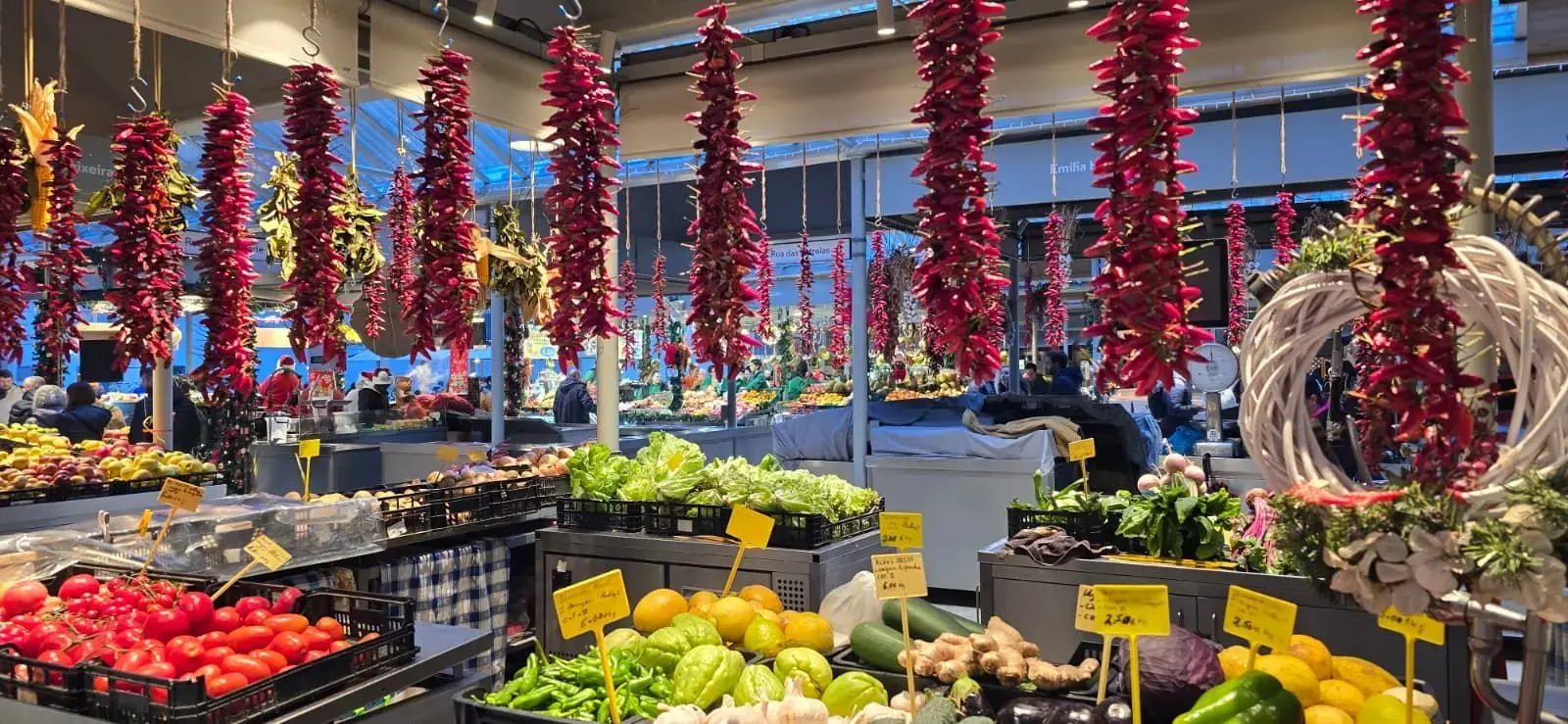 Strings of red chili peppers hanging over crates of fruit and vegetables in a busy indoor market, likely Lisbon, Portugal.