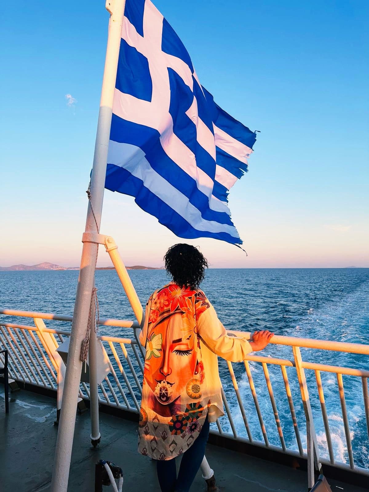 Greek flag flying above a traveler leaning on a ferry railing, looking over the Aegean Sea toward distant Greek islands, Greece.