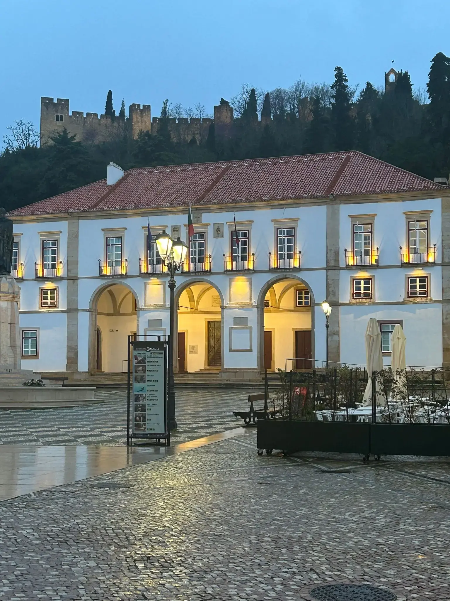 Leiria Castle on a tree-covered hill above a lit white building with arched loggia, evening in Leiria, Portugal