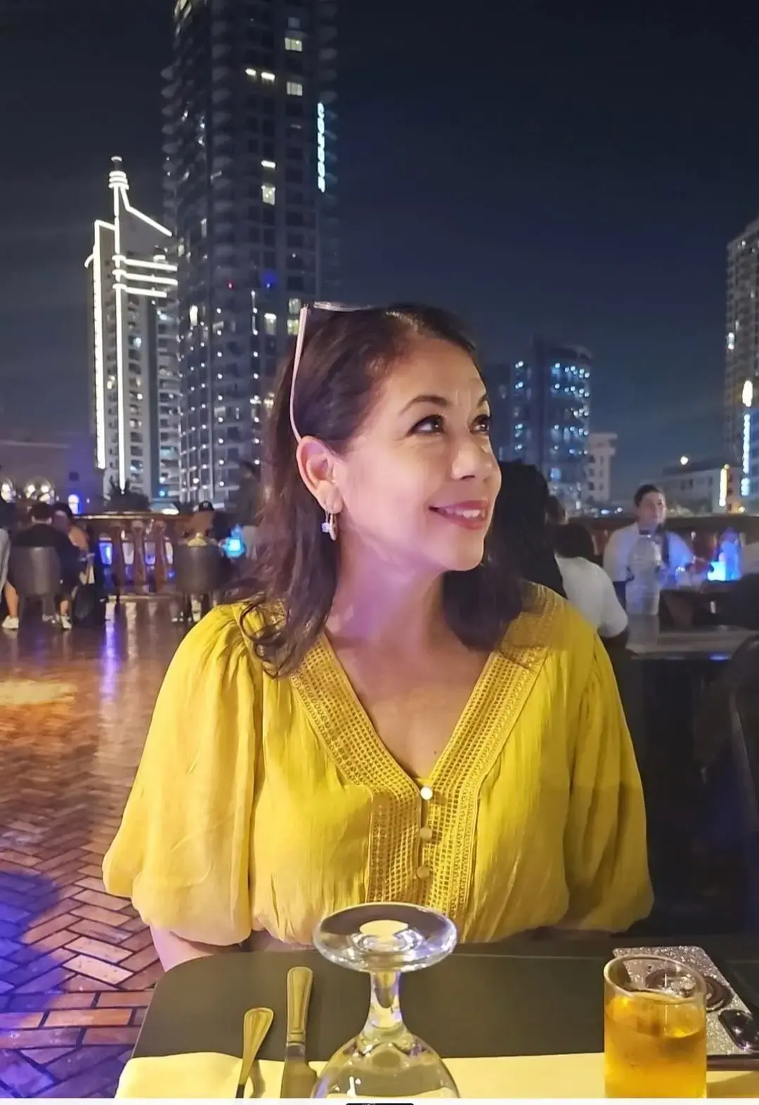 Woman at a rooftop restaurant looking up with illuminated Dubai skyline behind her, Dubai, United Arab Emirates.