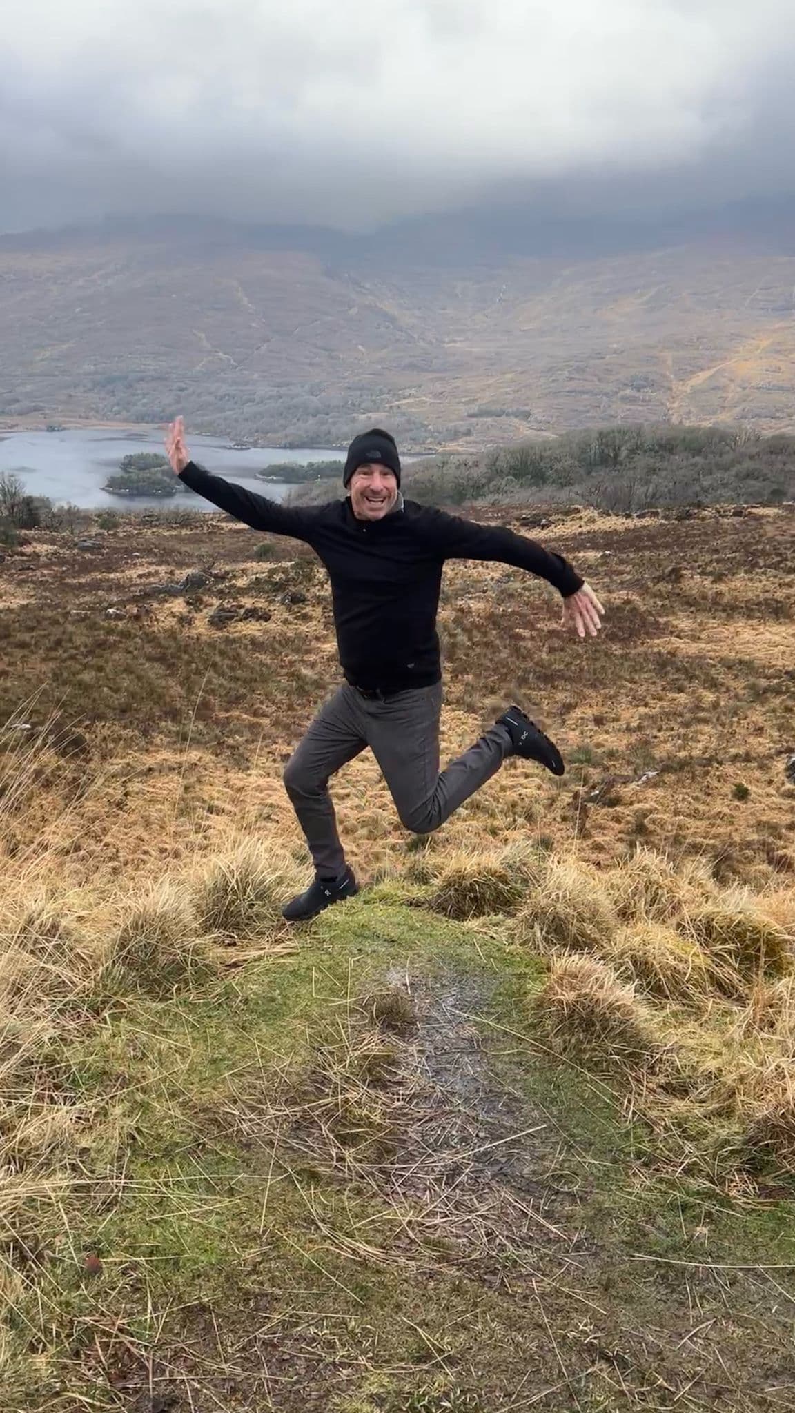 A man jumping on a grassy hill overlooking a lake and mountains in rural Ireland.