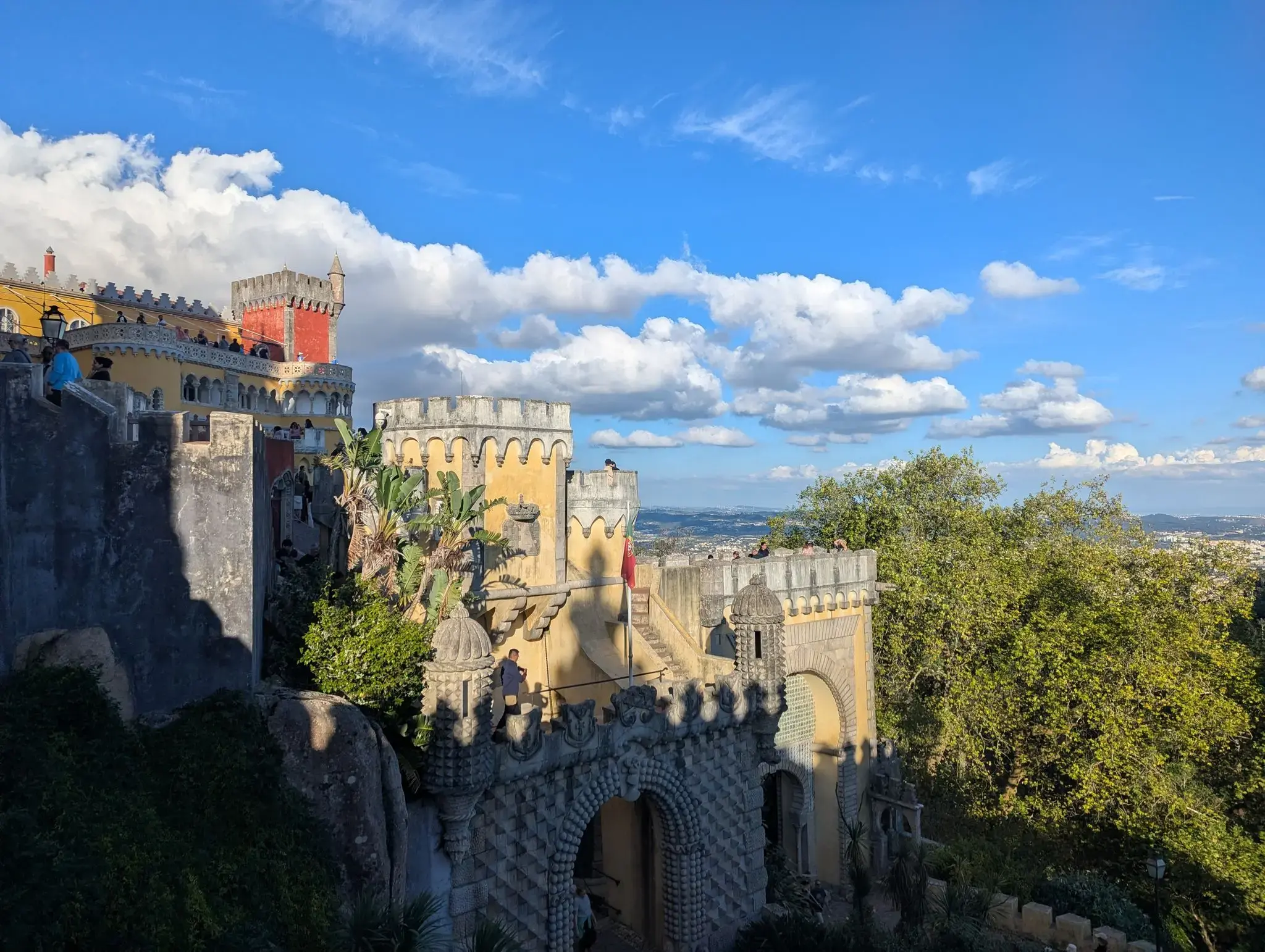 Pena Palace terraces and battlements in Sintra, Portugal, with visitors on a viewing terrace under a bright blue sky.