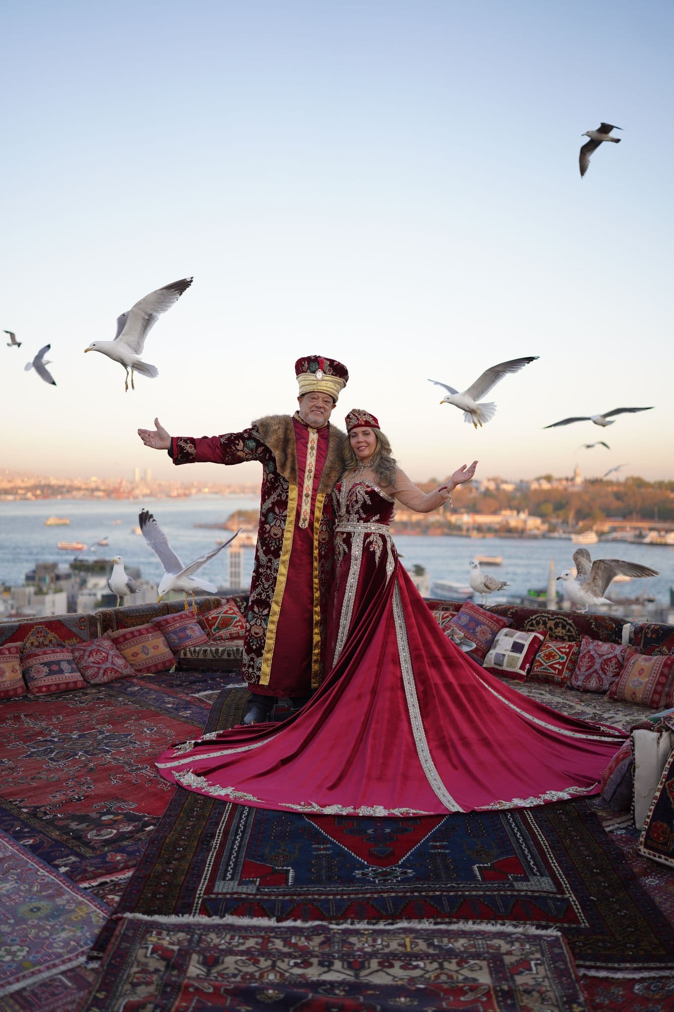 A couple in ornate red traditional costumes on a carpeted rooftop with seagulls over the Bosphorus, Galata, Istanbul, Turkey.