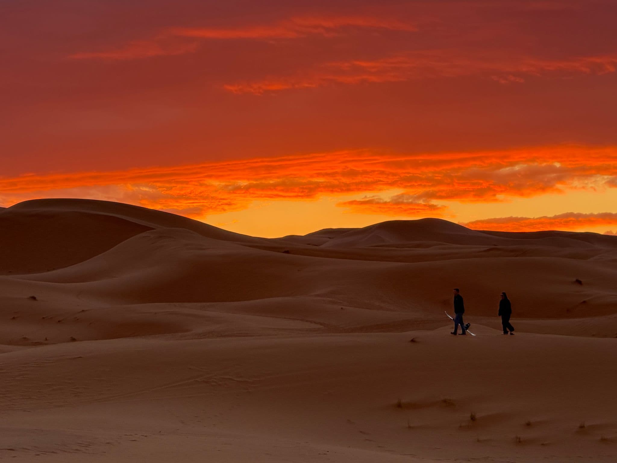 Sahara Desert dunes at sunrise near Erfoud, Morocco, two people walking across the sand carrying sandboards.