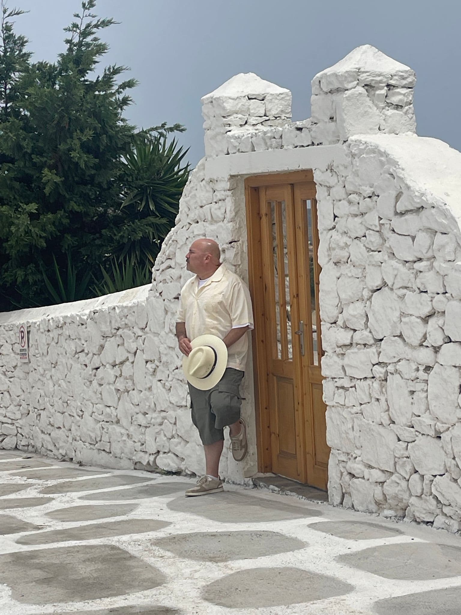 Santorini whitewashed doorway with a man leaning against the stone wall holding a straw hat on a paved street, Greece.