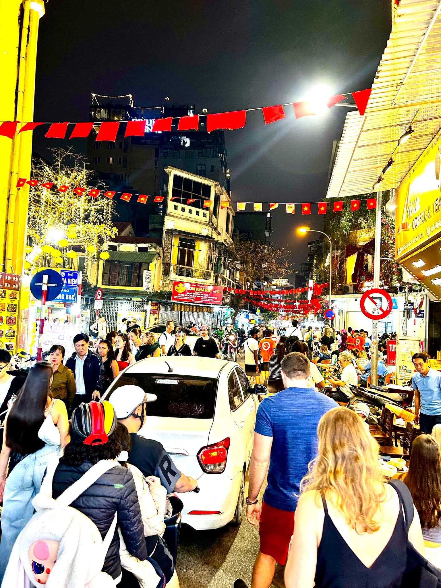 Crowded Ta Hien Street in Hanoi, Vietnam, with pedestrians, motorbikes, and red Vietnamese flags strung overhead at night.