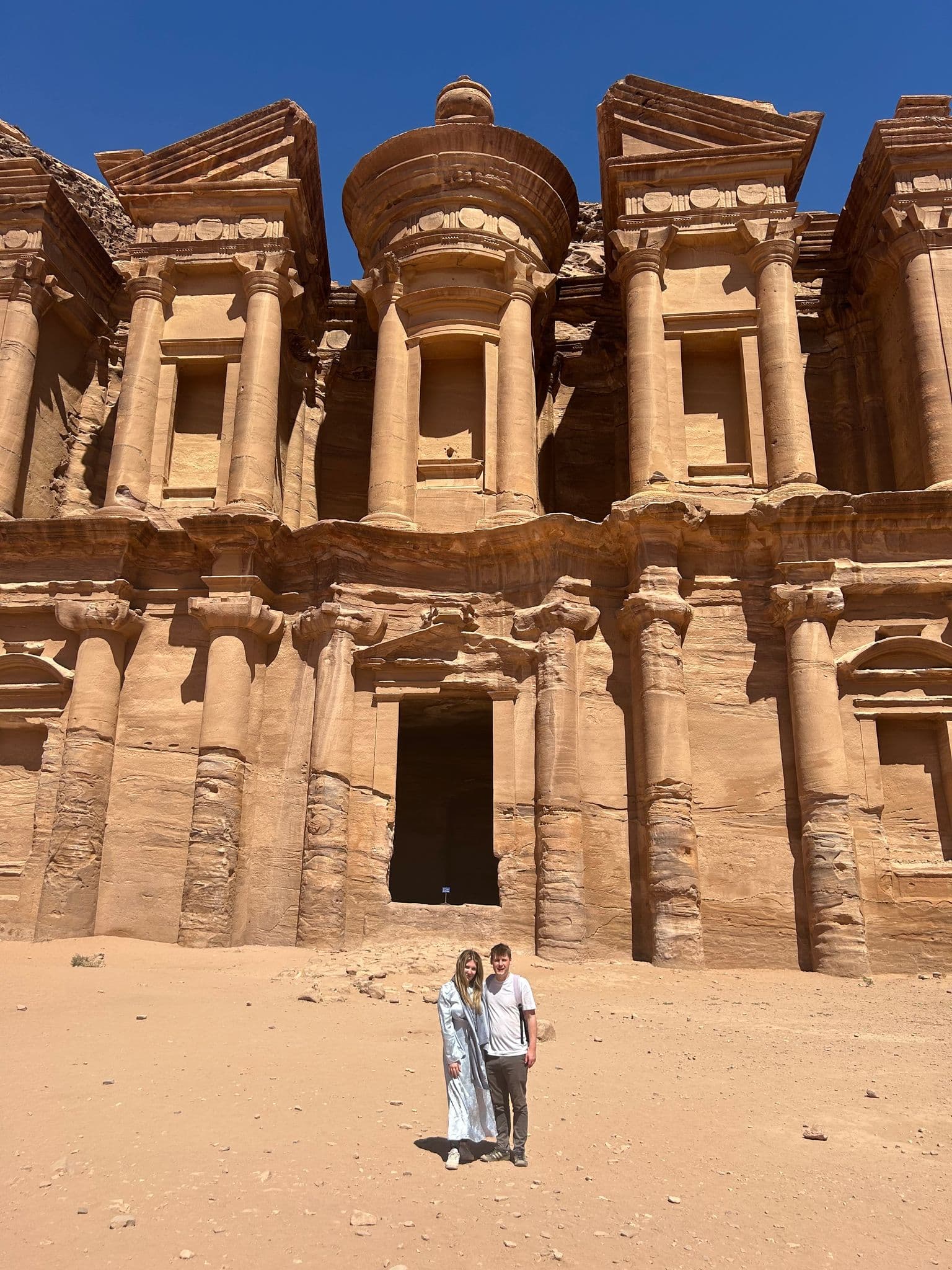 The Monastery (Ad Deir) carved into Petra's sandstone cliffs with two travelers standing in front, Jordan