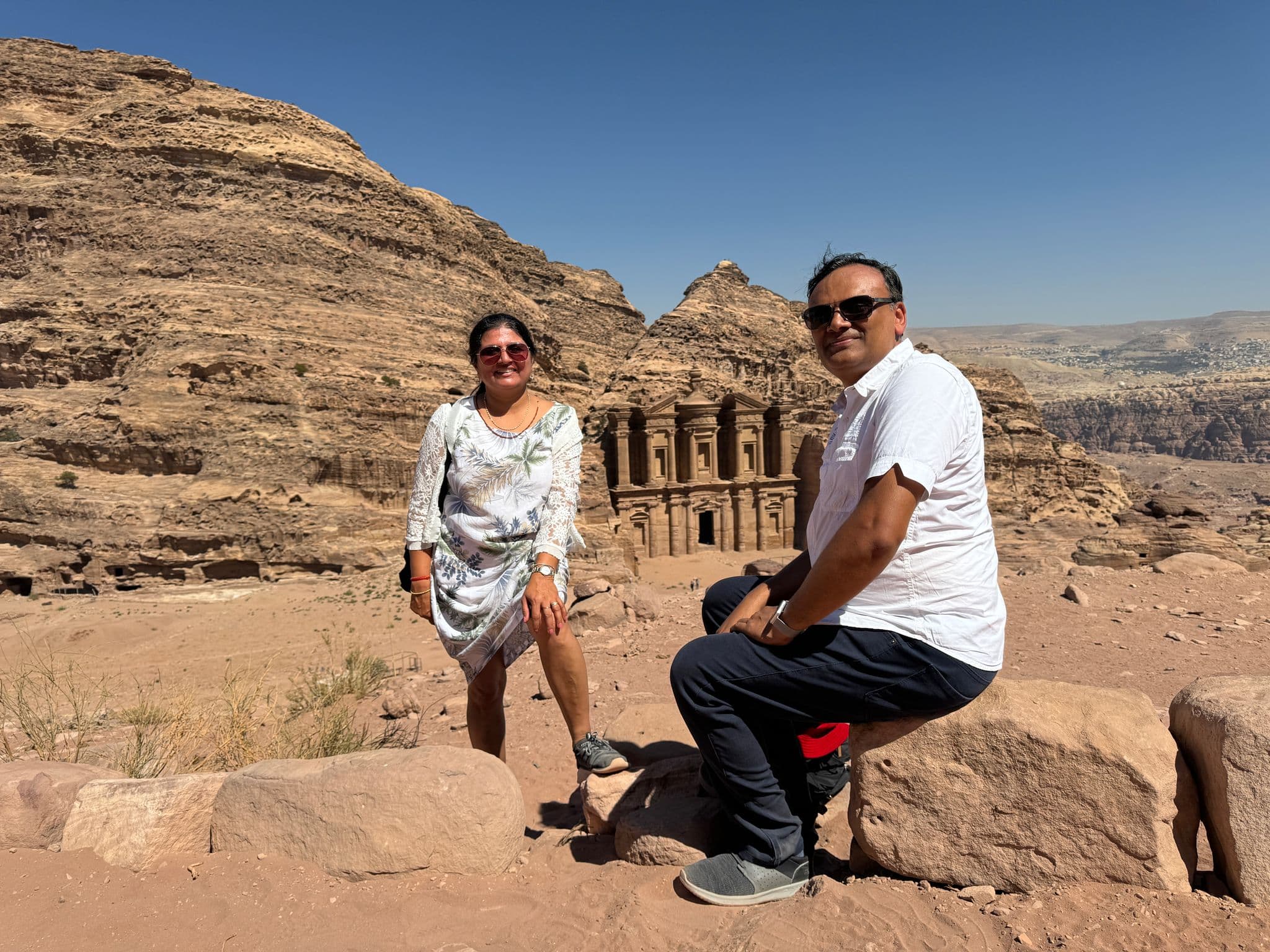 The Monastery (Ad Deir) at Petra, Jordan, carved into sandstone with two travelers posing on rocks in front on a sunny day.