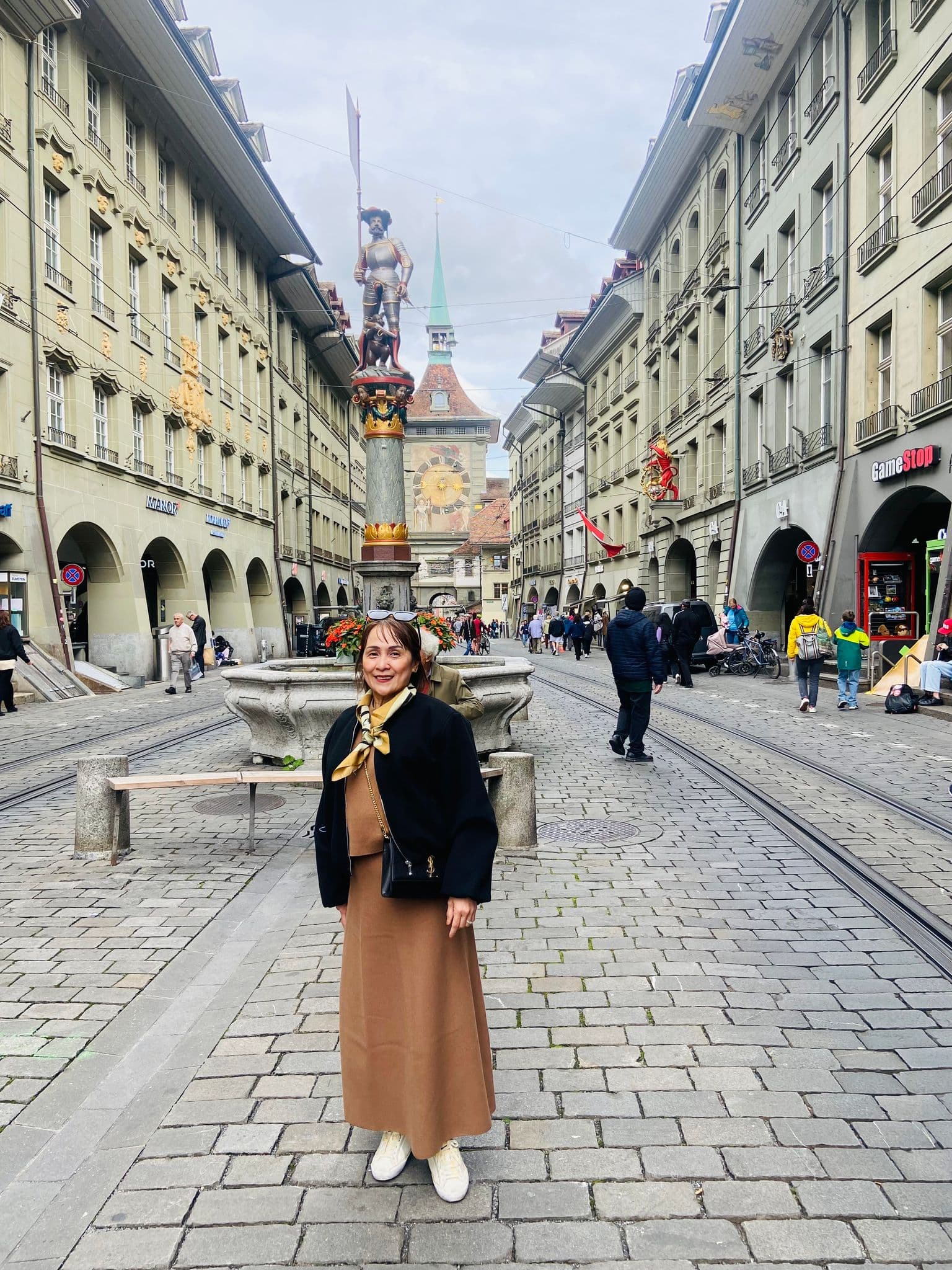 Zytglogge clock tower and medieval street in Bern, Switzerland, with a traveler posing by a fountain on cobbled tram tracks.