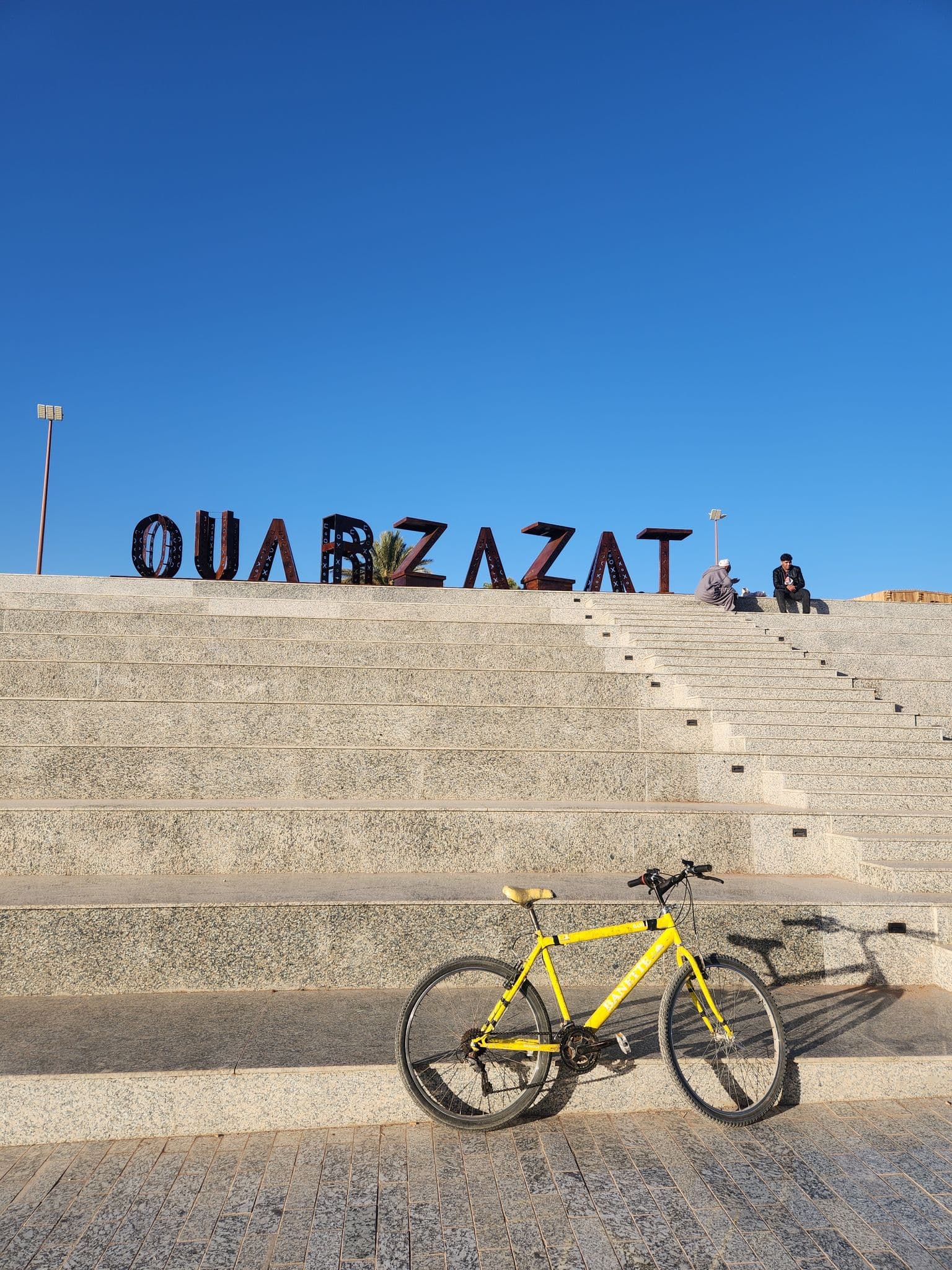 OUARZAZAT sign on stone steps with a yellow bicycle parked on the stairs and two people sitting near the top, Ouarzazat, Morocco.