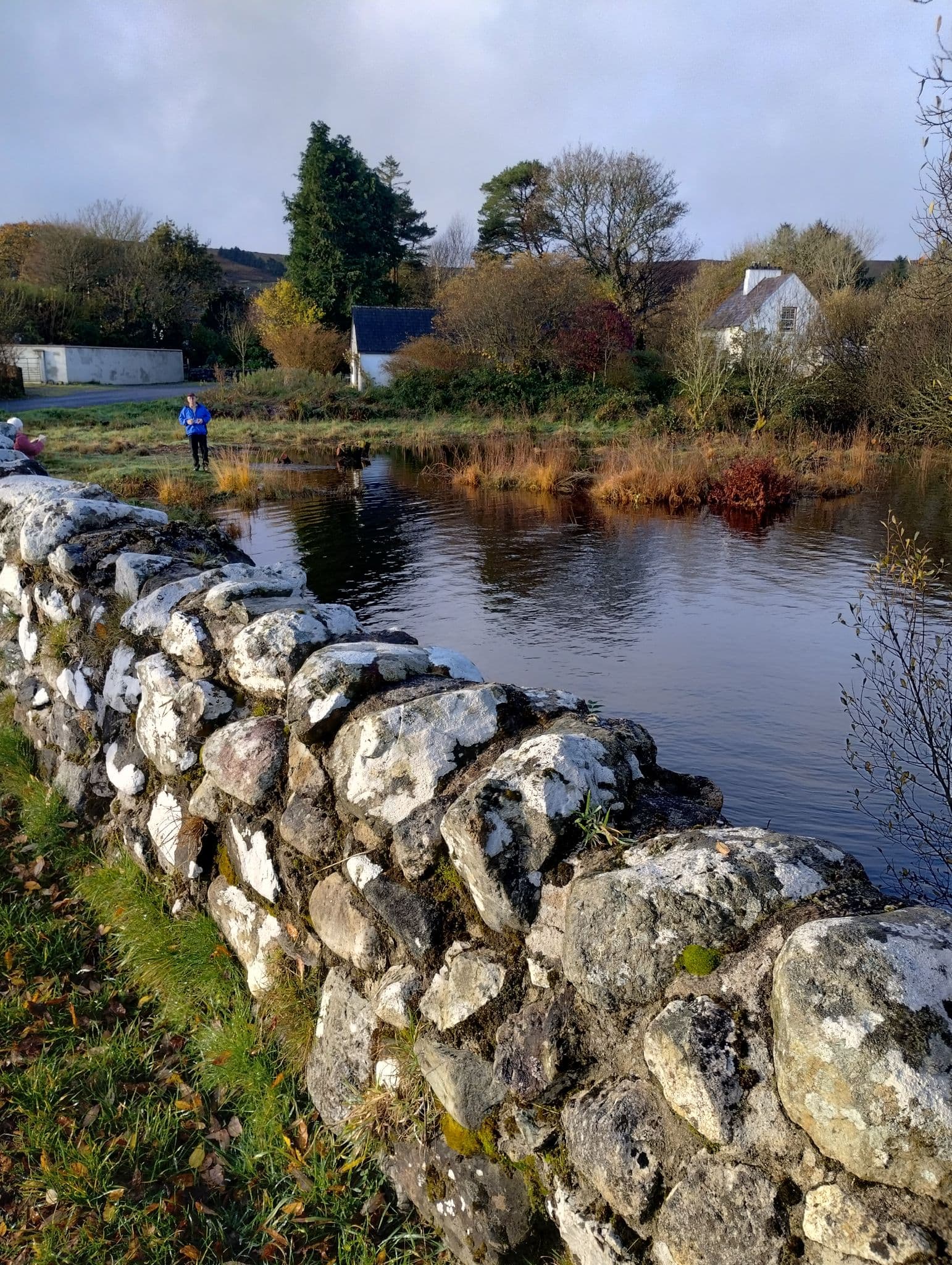 Stone wall and river at The Quiet Man filming location in Cong, Ireland, with a person standing on the far bank near white cottages.