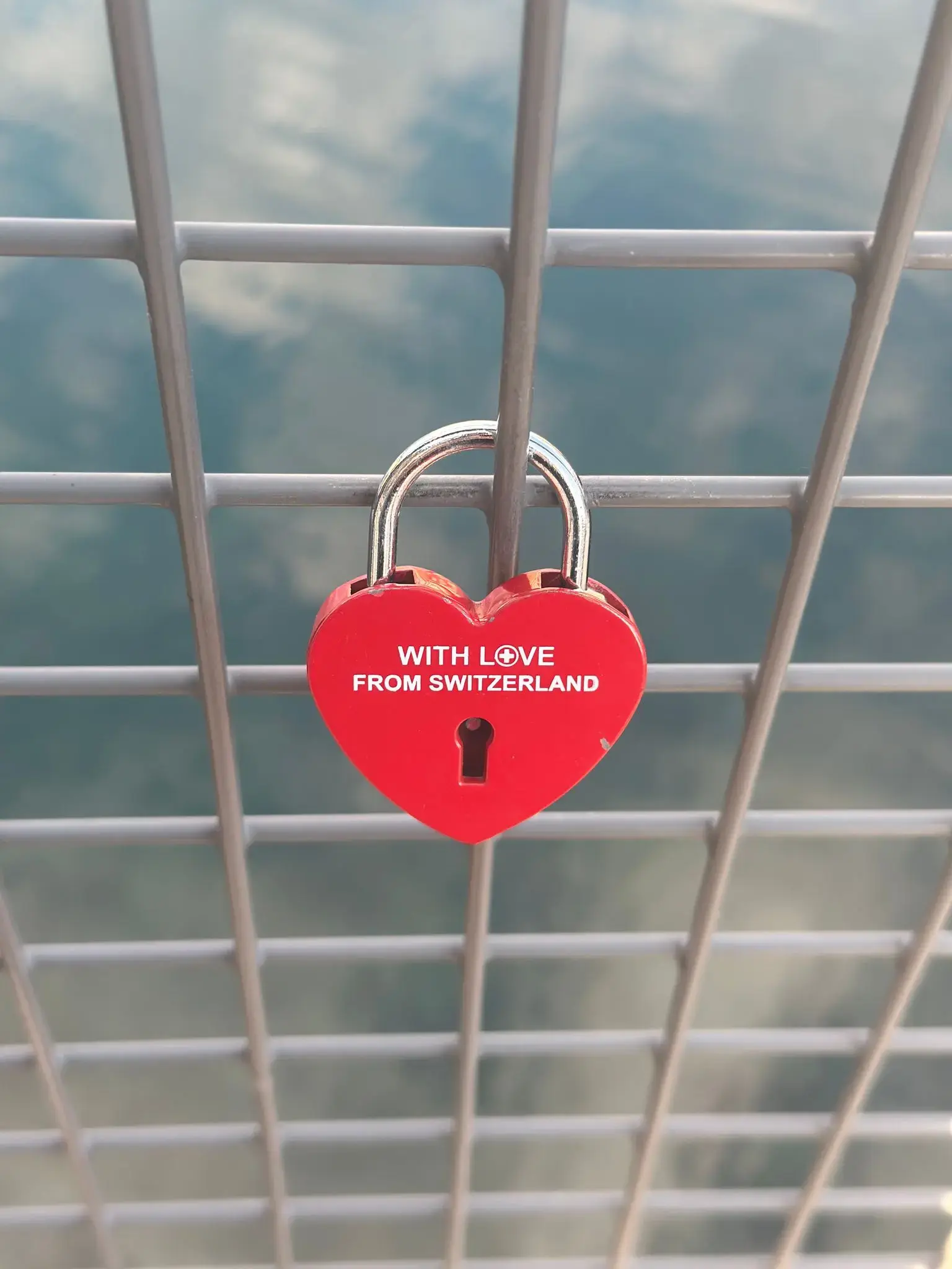 Red heart-shaped padlock reading "WITH LOVE FROM SWITZERLAND" locked to a metal bridge fence with water visible below, Switzerland.