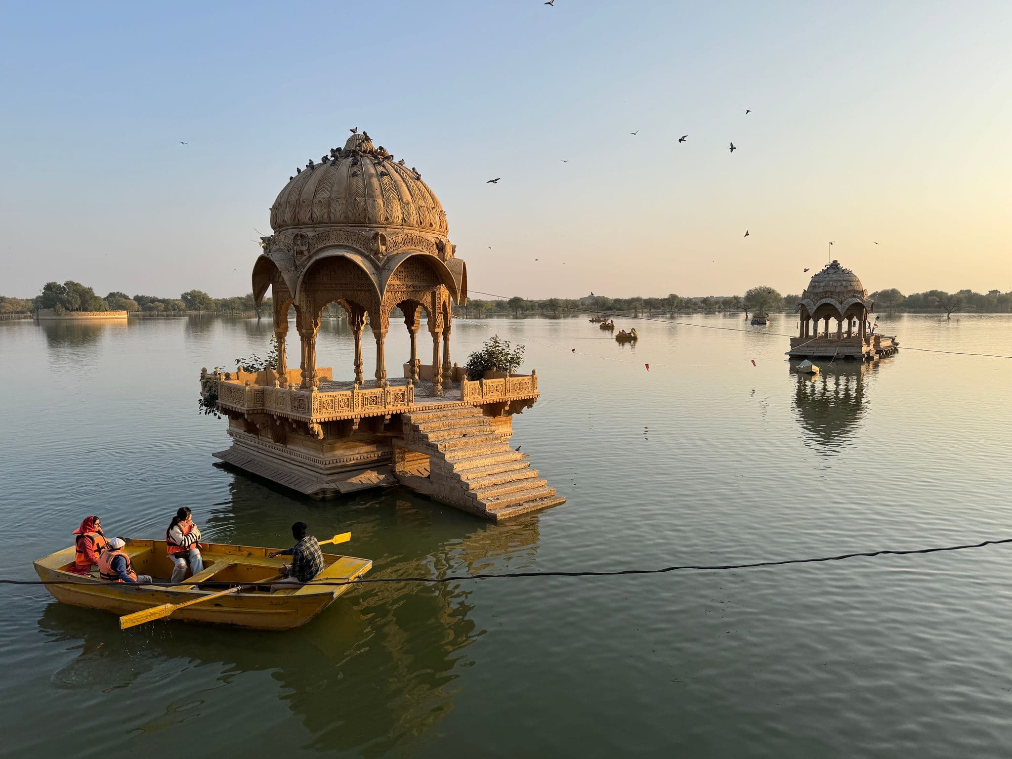 Ornate lakeside pavilion on Gadisar Lake in Jaisalmer, India, with a small rowboat of travelers passing in the foreground.