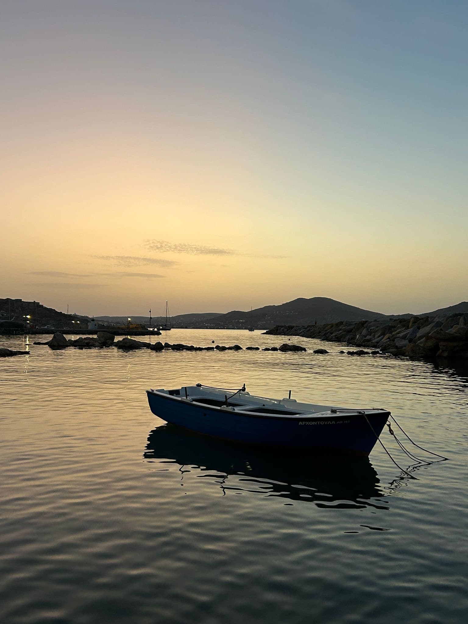 Small blue rowboat floating in a calm harbor at sunset with rocky shoreline and the hills of Paros, Greece.