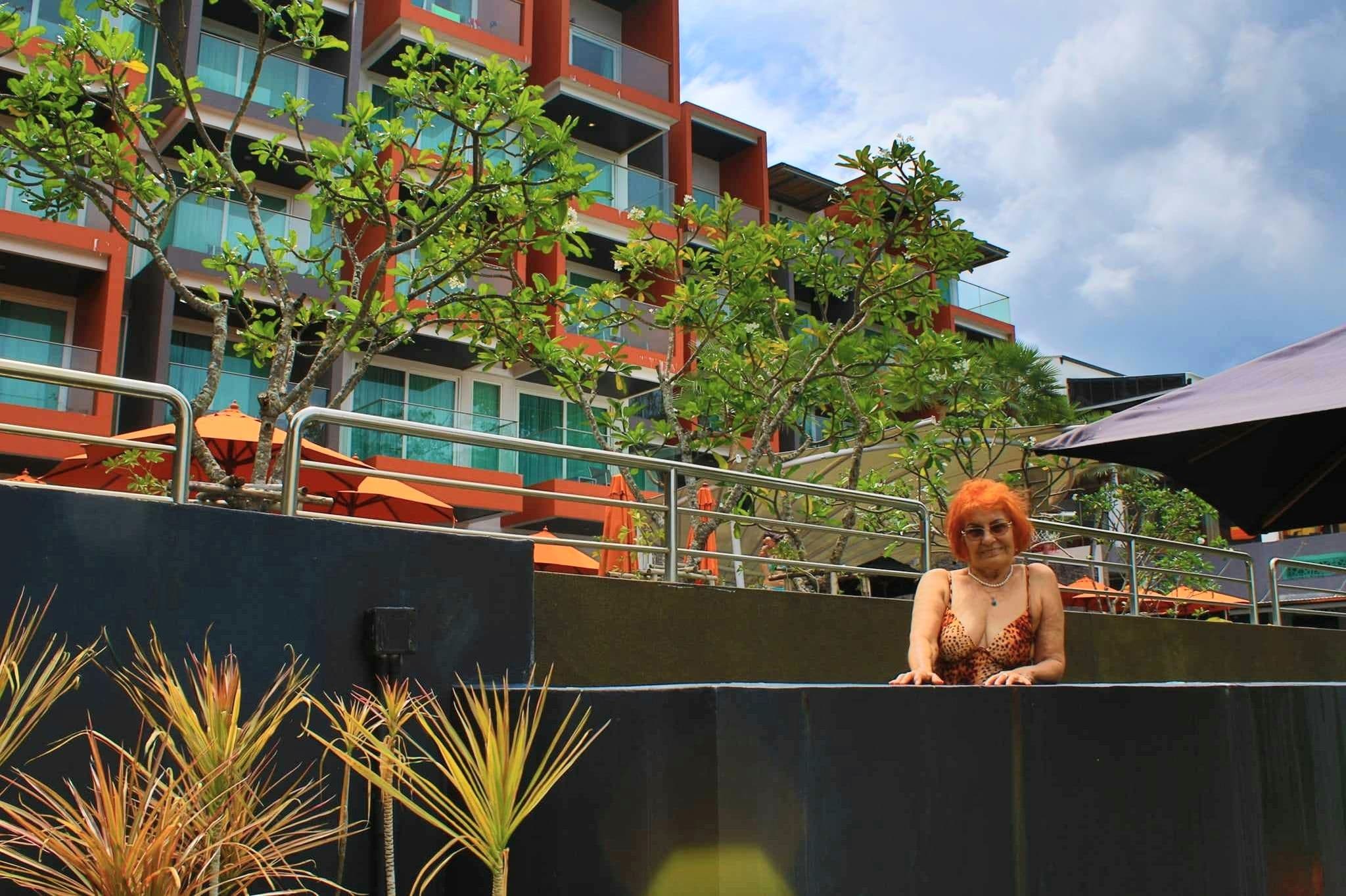Woman leaning on the edge of a hotel swimming pool with orange umbrellas and a multi-story resort building behind, Thailand.