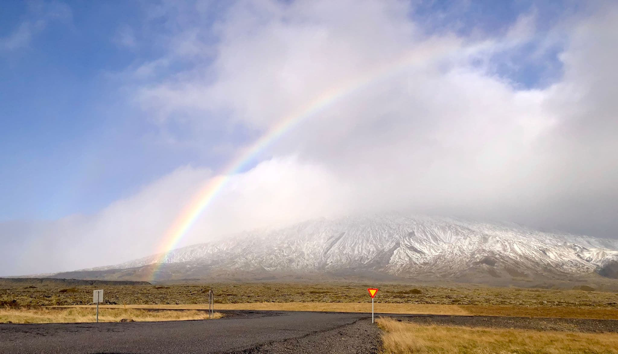 Rainbow arching over a snow-dusted mountain and mossy lava fields with a roadside yield sign in Iceland.