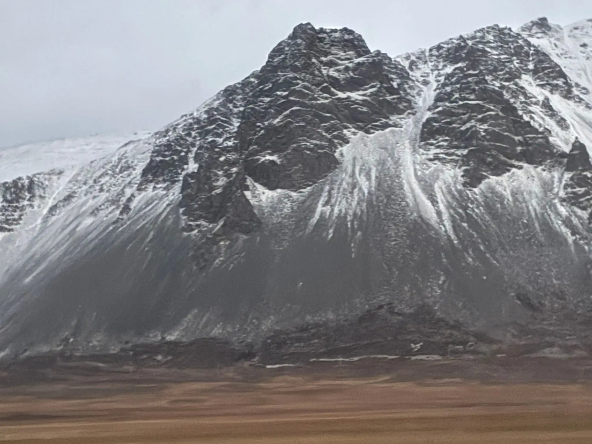 Snow-dusted rocky mountain rising above a flat brown plain under a pale morning sky.