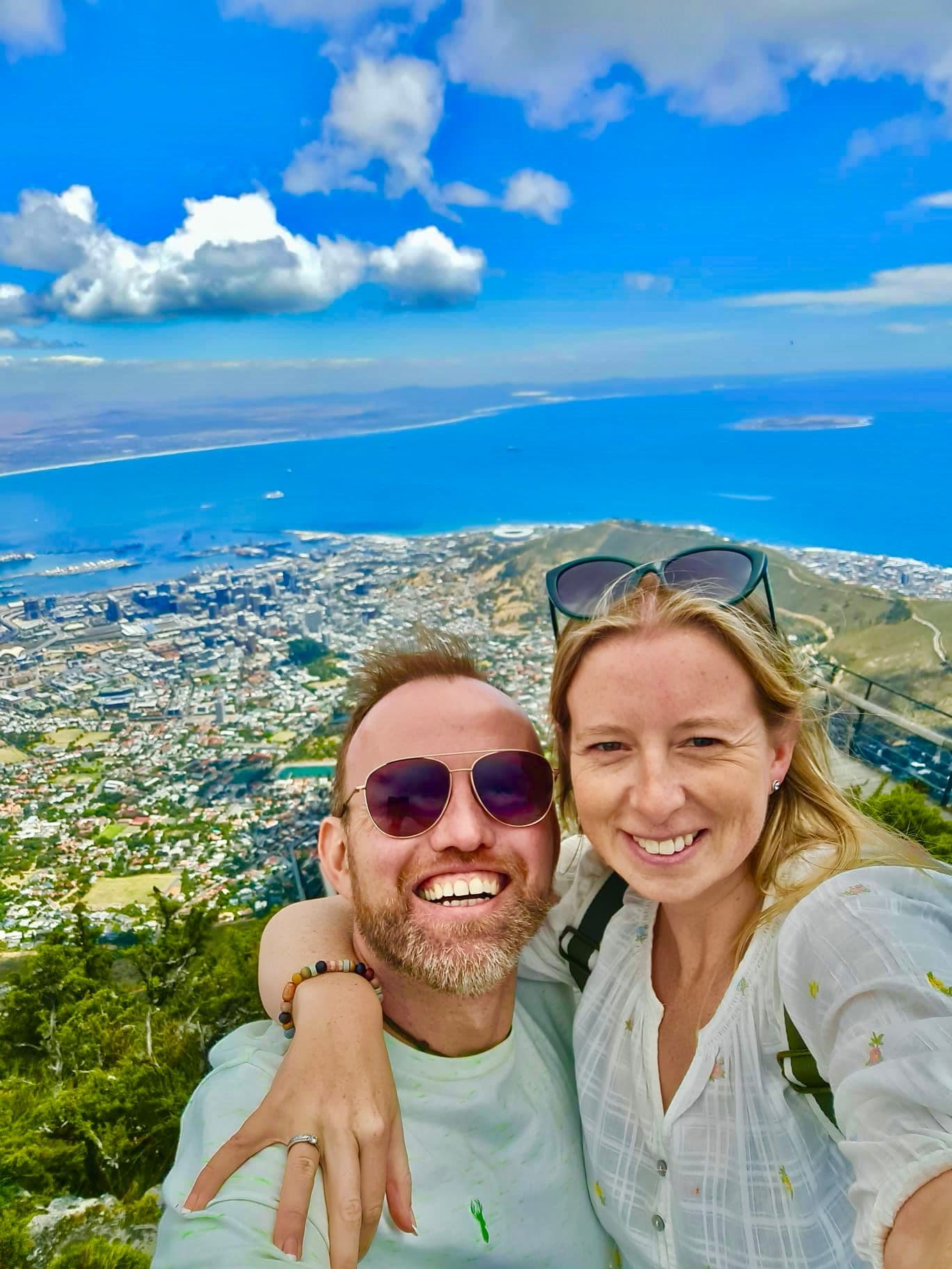 Table Mountain summit overlooking Cape Town, South Africa, with two travelers taking a selfie showing the city and ocean below.