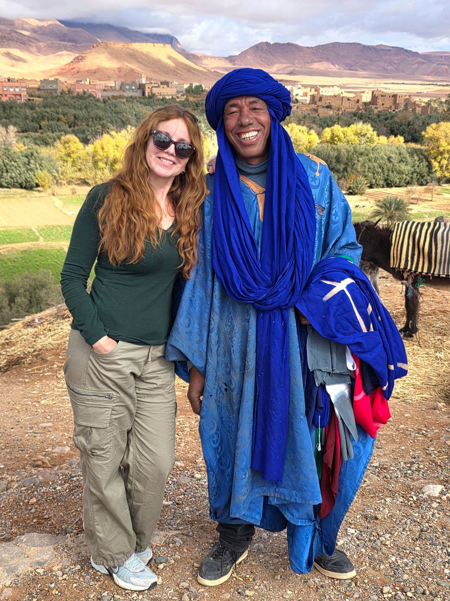 Man in vibrant blue robe standing with a traveler before a palm oasis and kasbahs in the Draa Valley, Morocco.