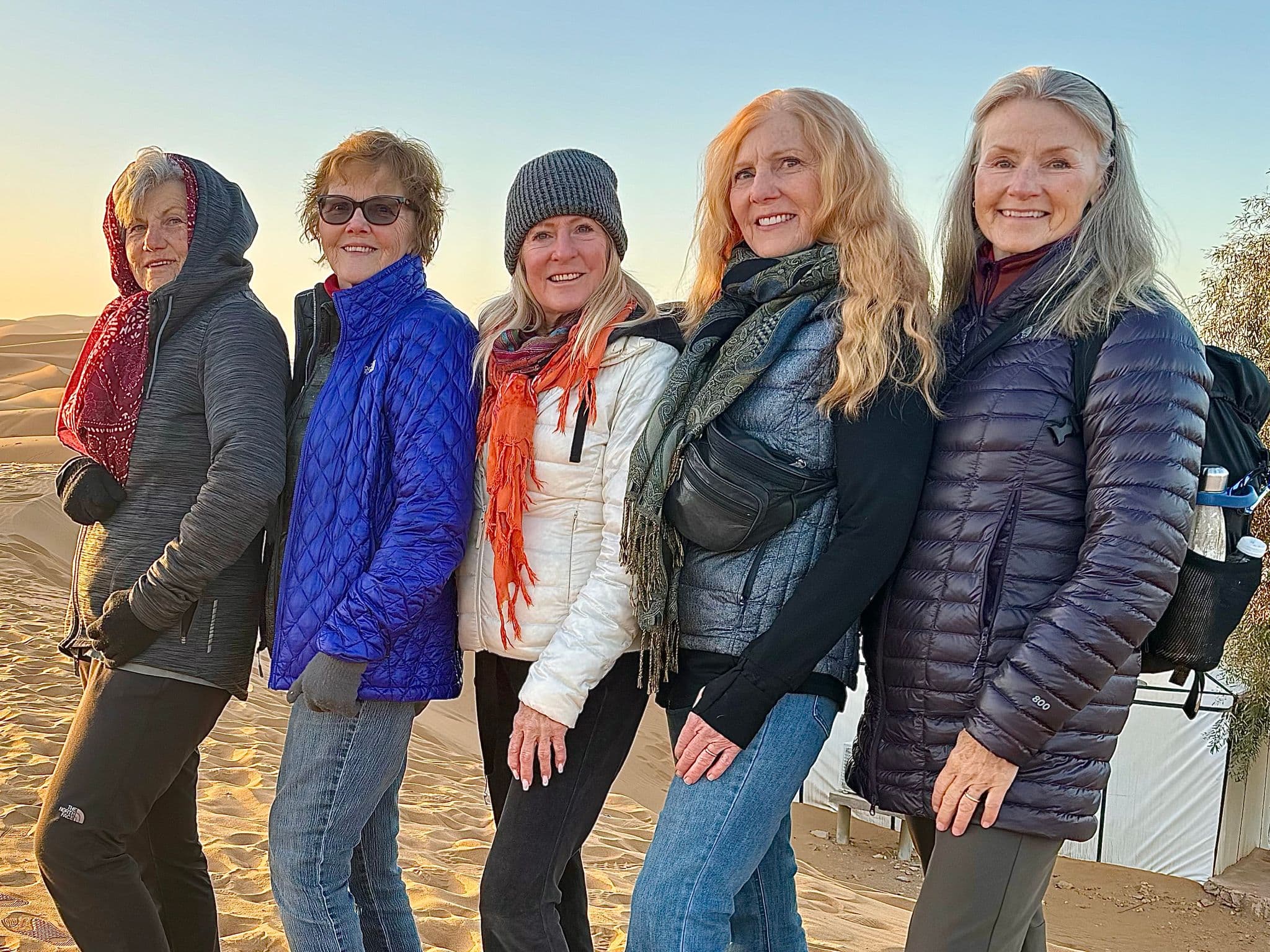 Five women posing together on sand dunes at a camp in the Sahara Desert.