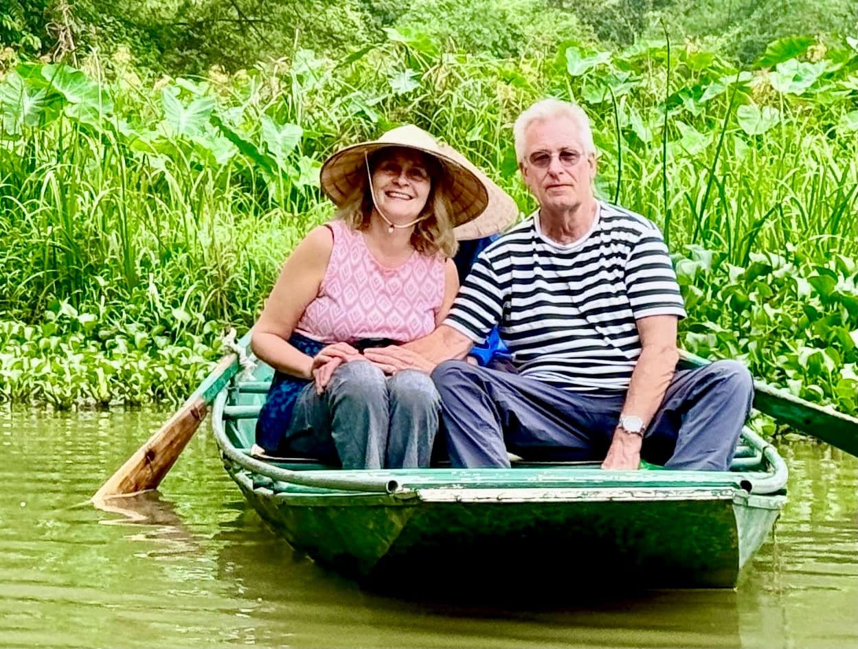Rowboat carrying two travelers through lush river vegetation in Ninh Binh National Park, near Hanoi, Vietnam.