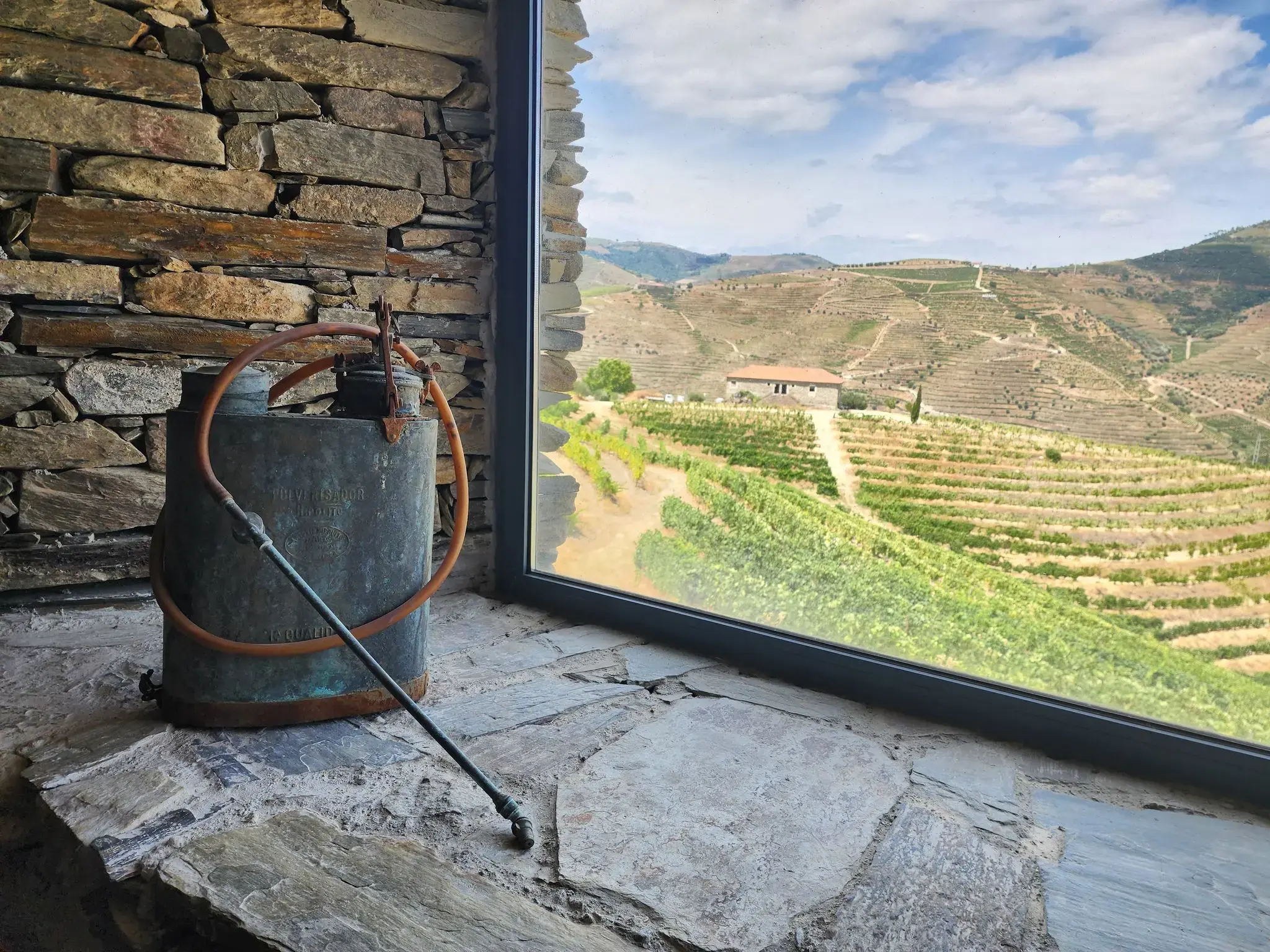 Terraced vineyards of the Douro Valley, Portugal, seen through a window with an old metal sprayer on a stone windowsill.