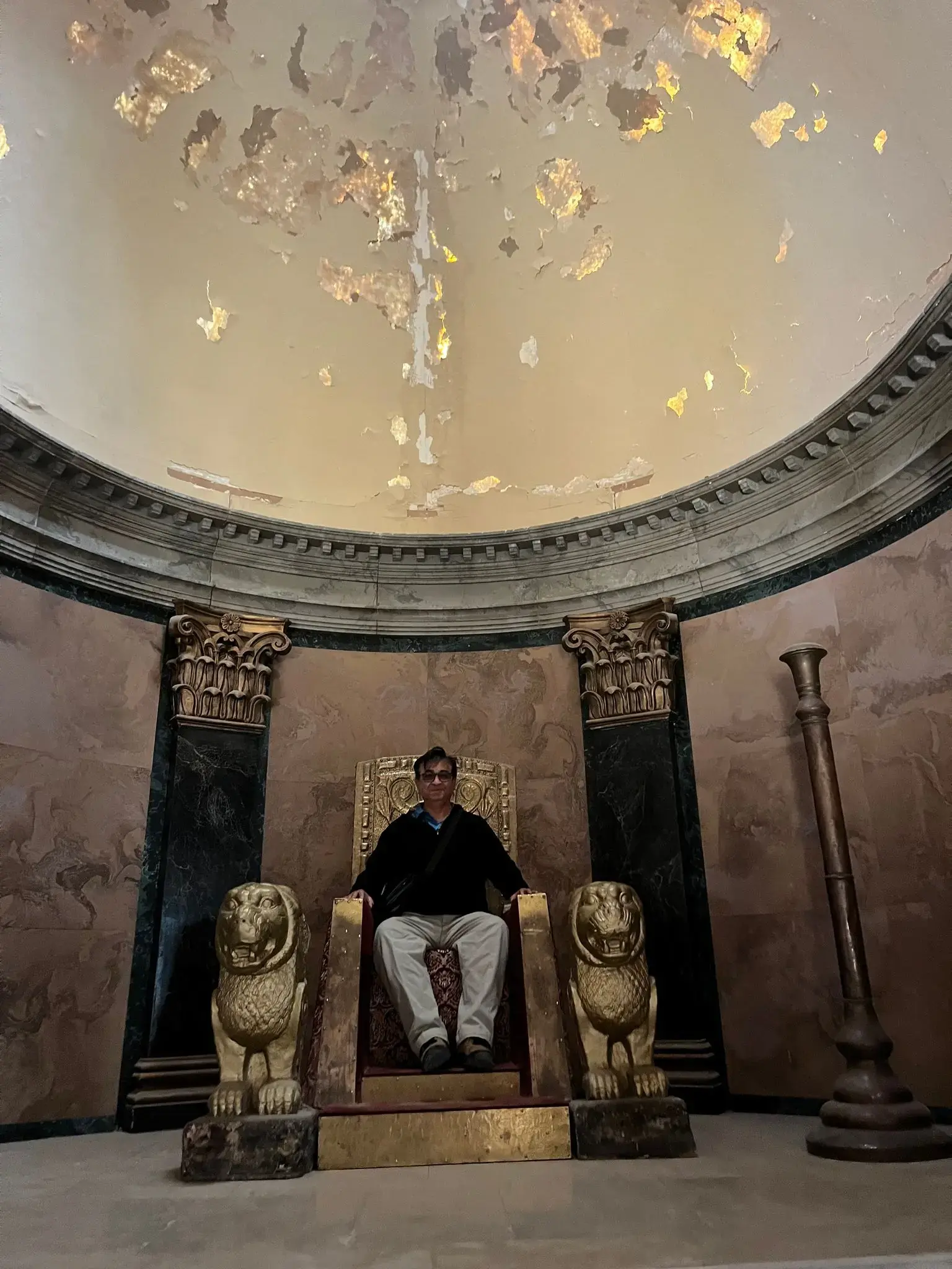 Gilded throne with carved lion armrests and a person seated beneath a peeling domed ceiling in Morocco.