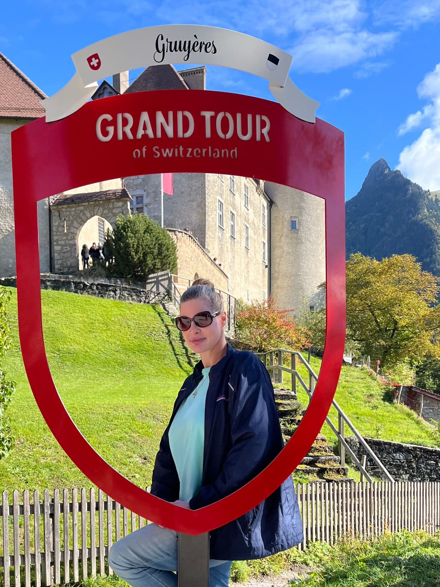 Red "Grand Tour of Switzerland" sign framing a woman posing in front of Château de Gruyères, Gruyères, Switzerland.