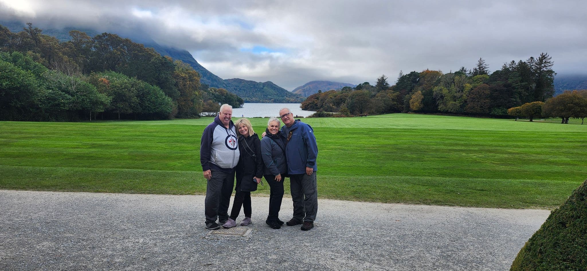 Muckross Lake in Killarney National Park, Ireland, with four travelers standing on a path and green lawns in the foreground.