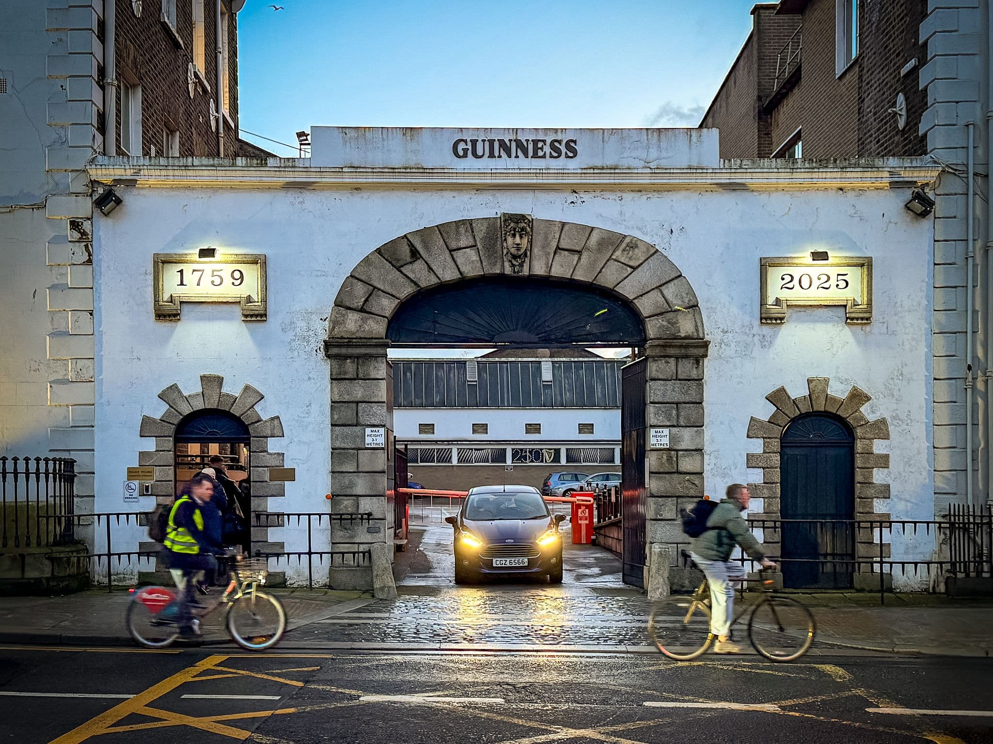Guinness brewery entrance at St. James's Gate, Dublin, Ireland, with a car passing through the arch and cyclists riding past on the street.