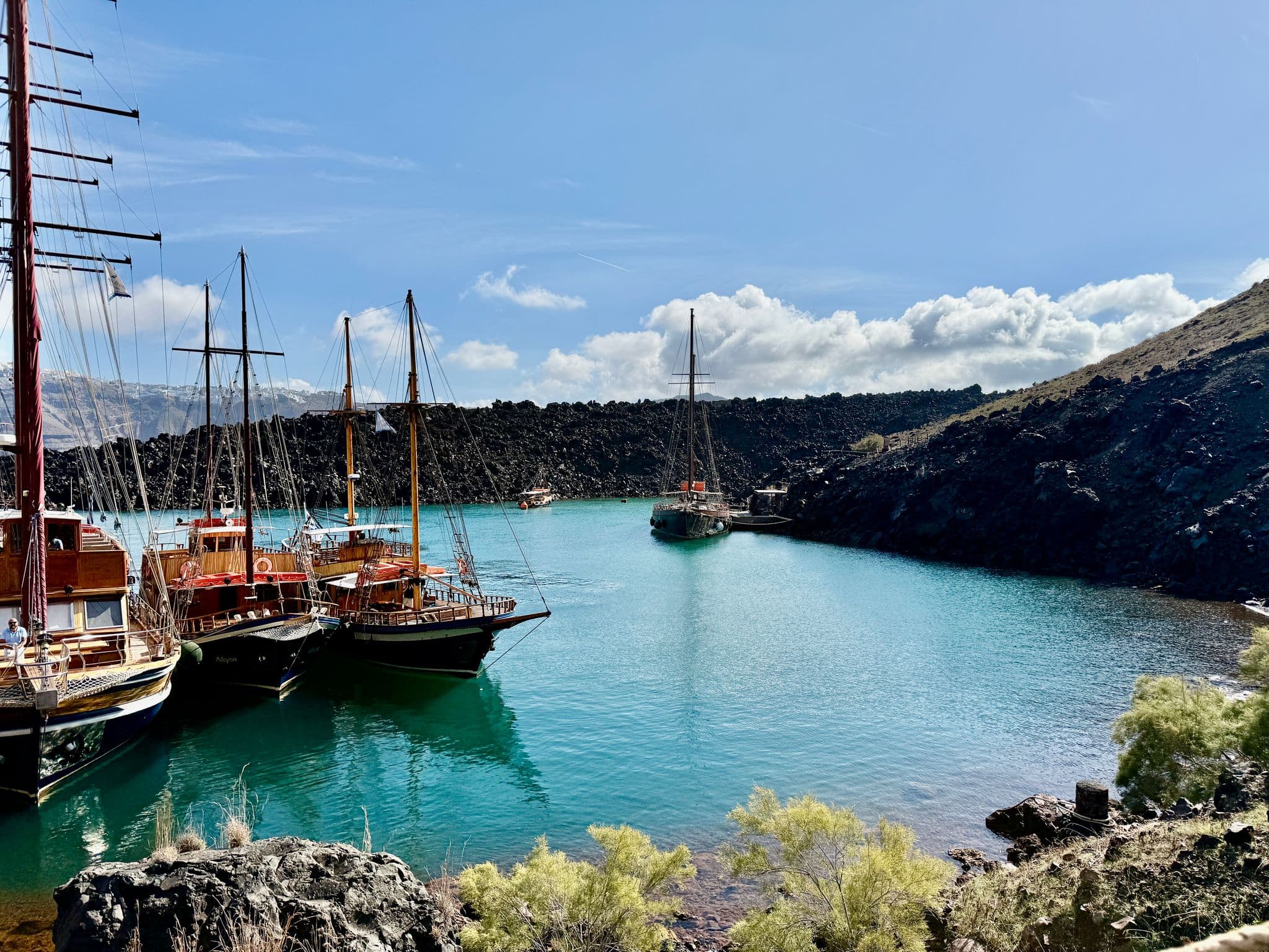 Santorini caldera with turquoise water and moored wooden sailboats beside volcanic cliffs on Thirassia, Greece.
