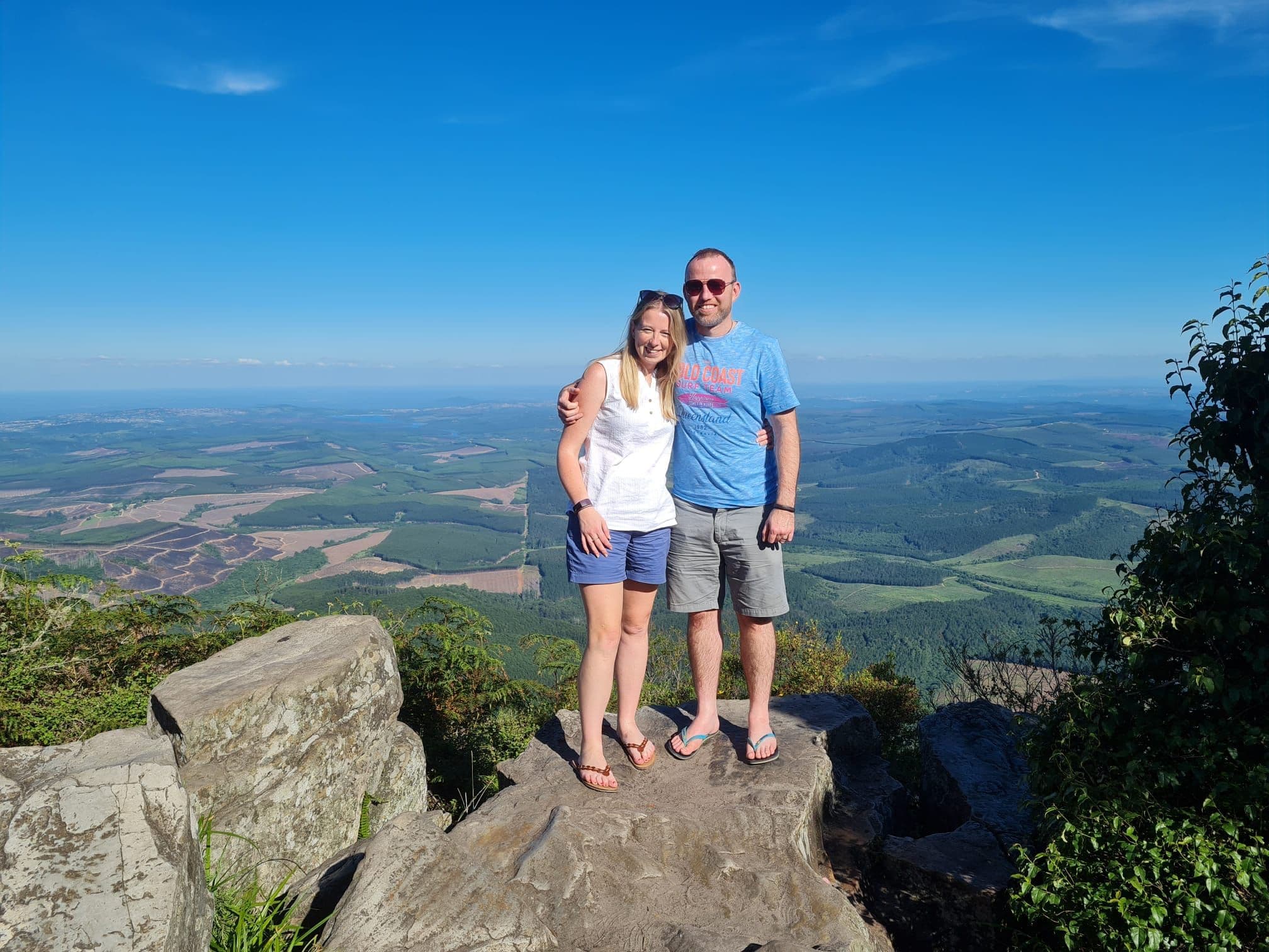 Two people standing on a rocky viewpoint overlooking rolling plains and distant coastline near Johannesburg, South Africa.