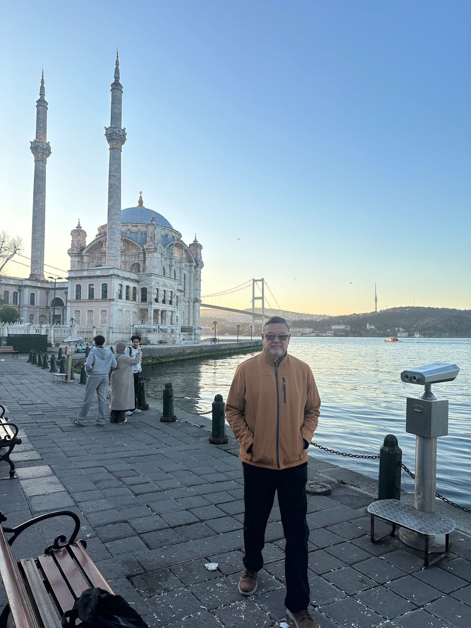 Ortaköy Mosque by the Bosphorus in Istanbul, Turkey, with a man standing on the waterside promenade and the Bosphorus Bridge in the background.