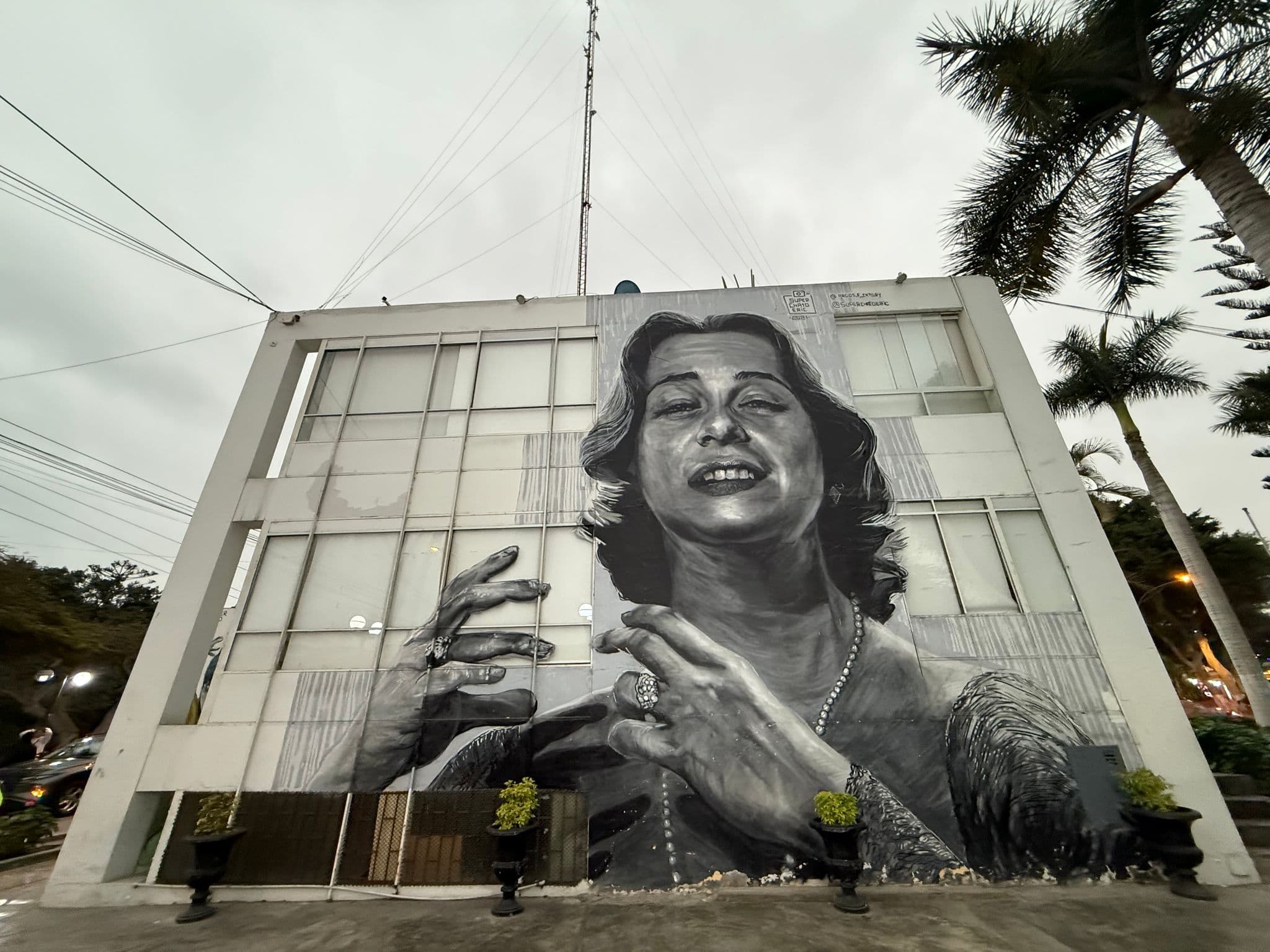 Large black-and-white mural of a woman covering a multi-story building facade, with palm trees on a street in Lima, Peru.