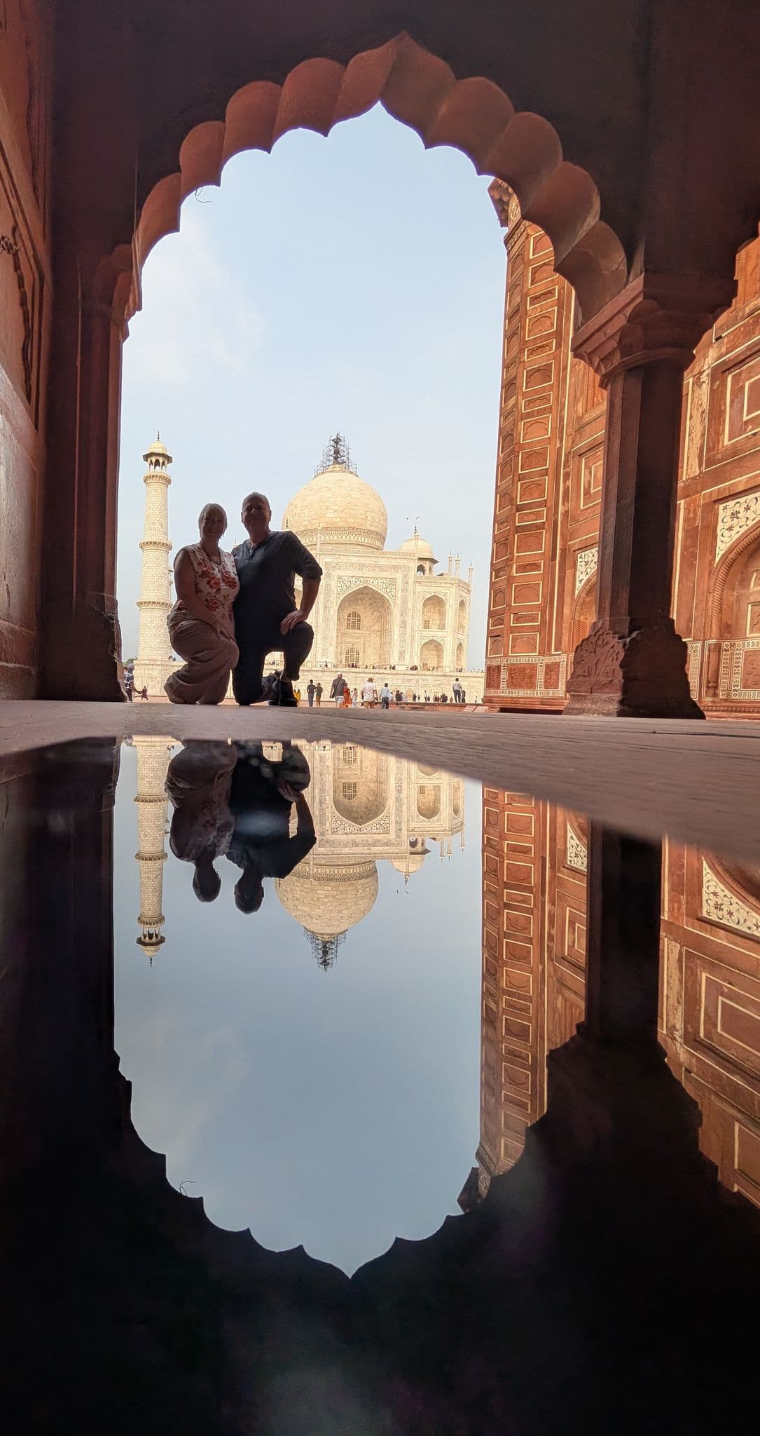 Taj Mahal framed by a red sandstone arch with two people kneeling in front and its reflection in a puddle, Agra, India.