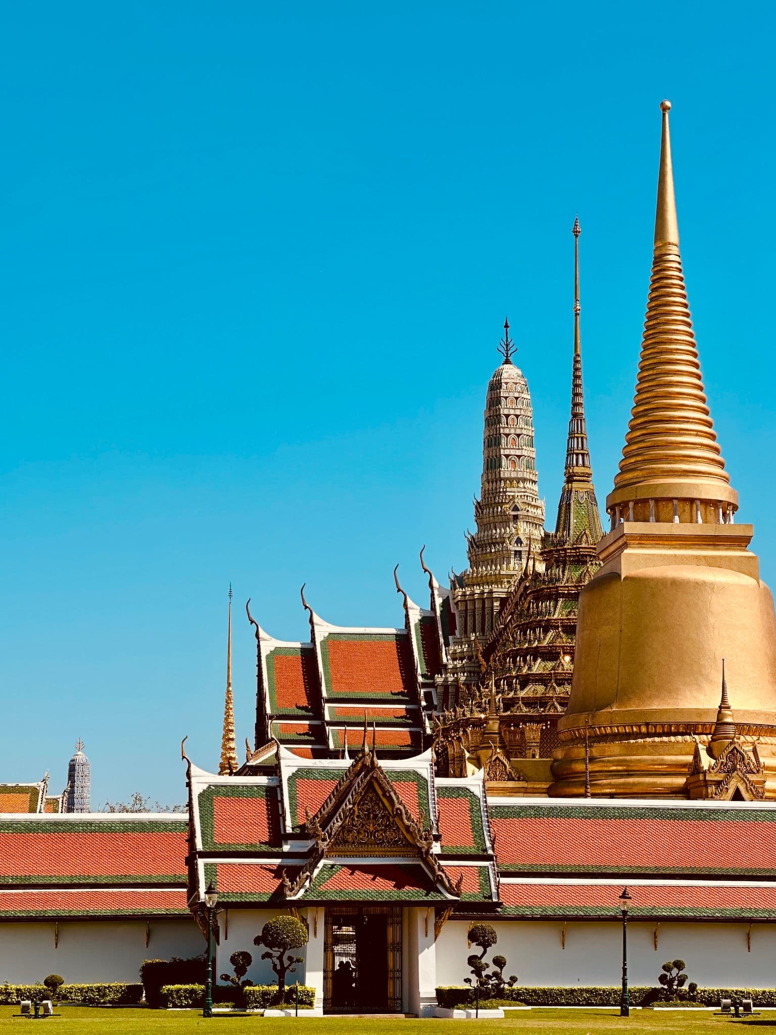 Golden stupa and ornate temple roofs of the Grand Palace and Wat Phra Kaew in Bangkok, Thailand against a clear blue sky.