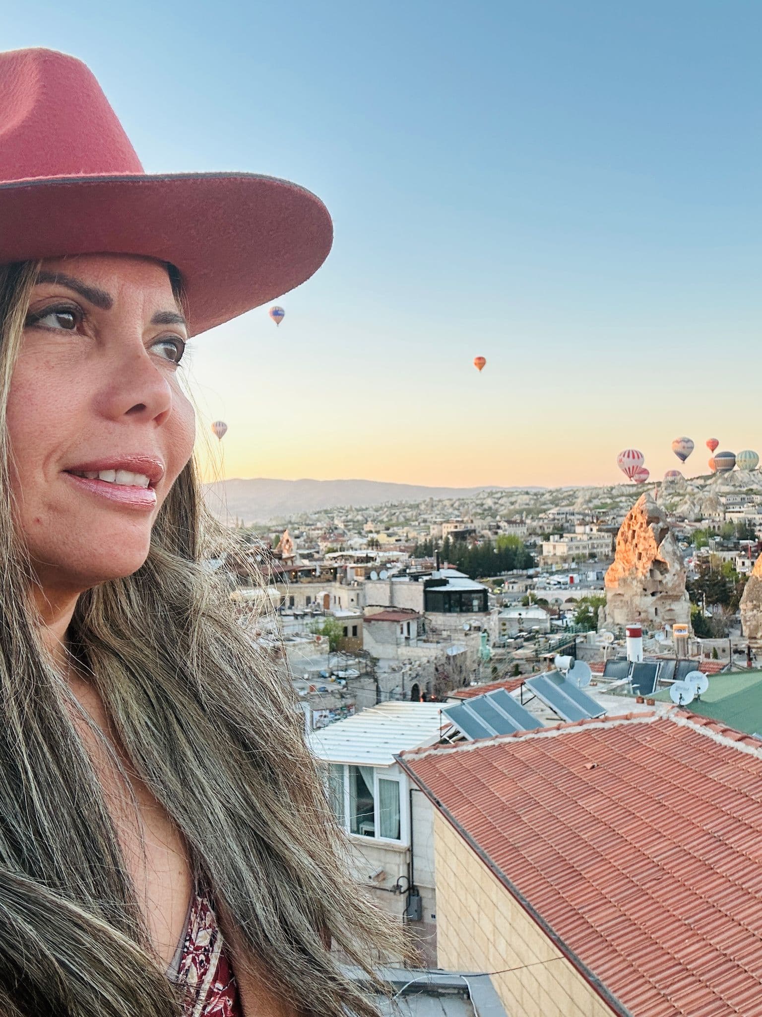 Hot air balloons over Göreme in Cappadocia, Turkey at sunrise with a woman in a hat on a rooftop.