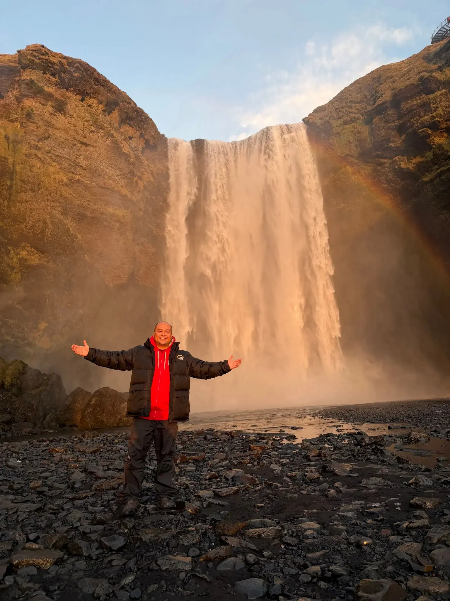 Skógafoss waterfall with a rainbow and a person standing on the rocky shore with arms outstretched, South Coast, Iceland