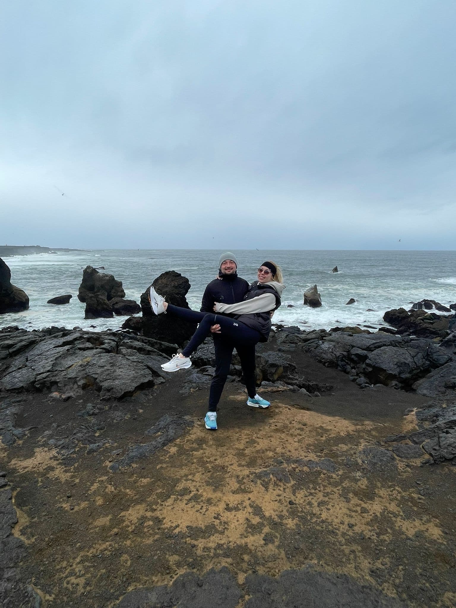 Couple at Valahnúkamöl on the rocky Iceland coast, man lifting his wife near the ocean.