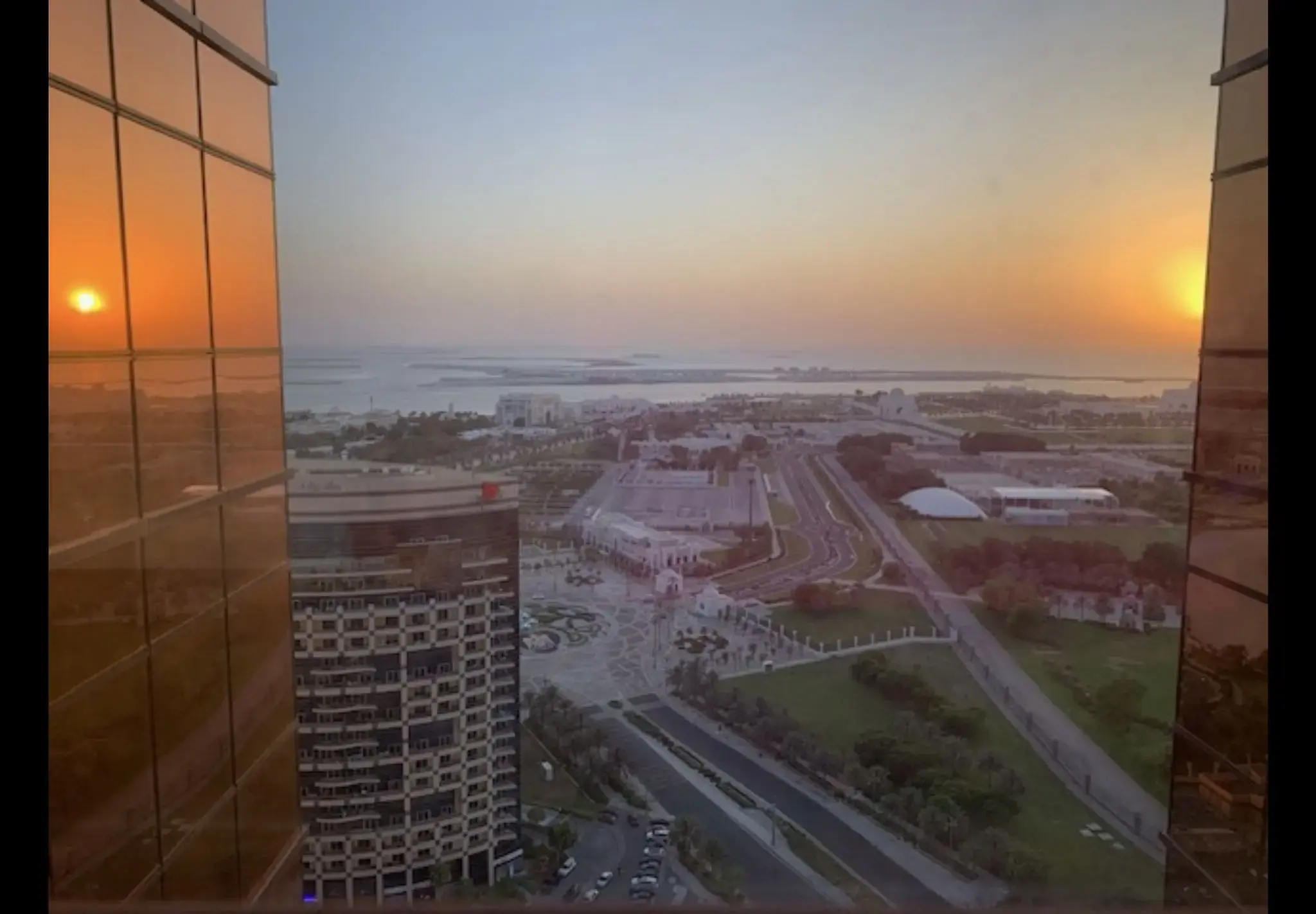 Sunset over a coastal city skyline seen through a high-rise glass window, Abu Dhabi, United Arab Emirates
